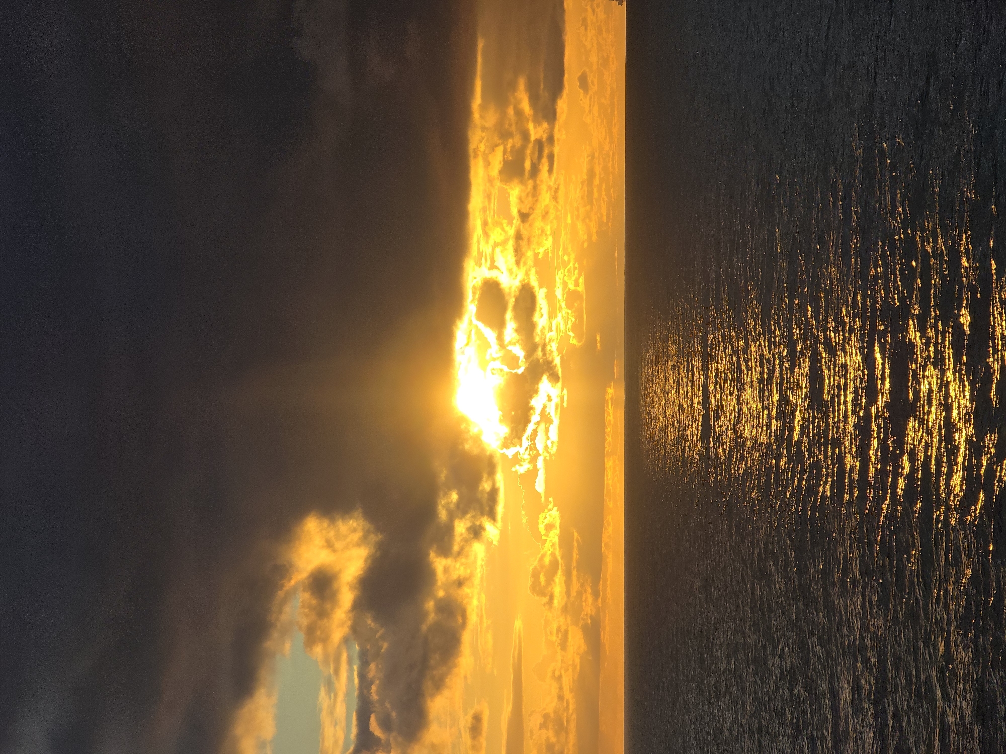 The image shows a sunset over water, possibly an ocean or large lake. The sun is setting behind clouds, creating a bright, golden light in the center of the image. The water reflects this light, creating a shimmering effect. The sky is dark on either side of the sun. The location is unknown.