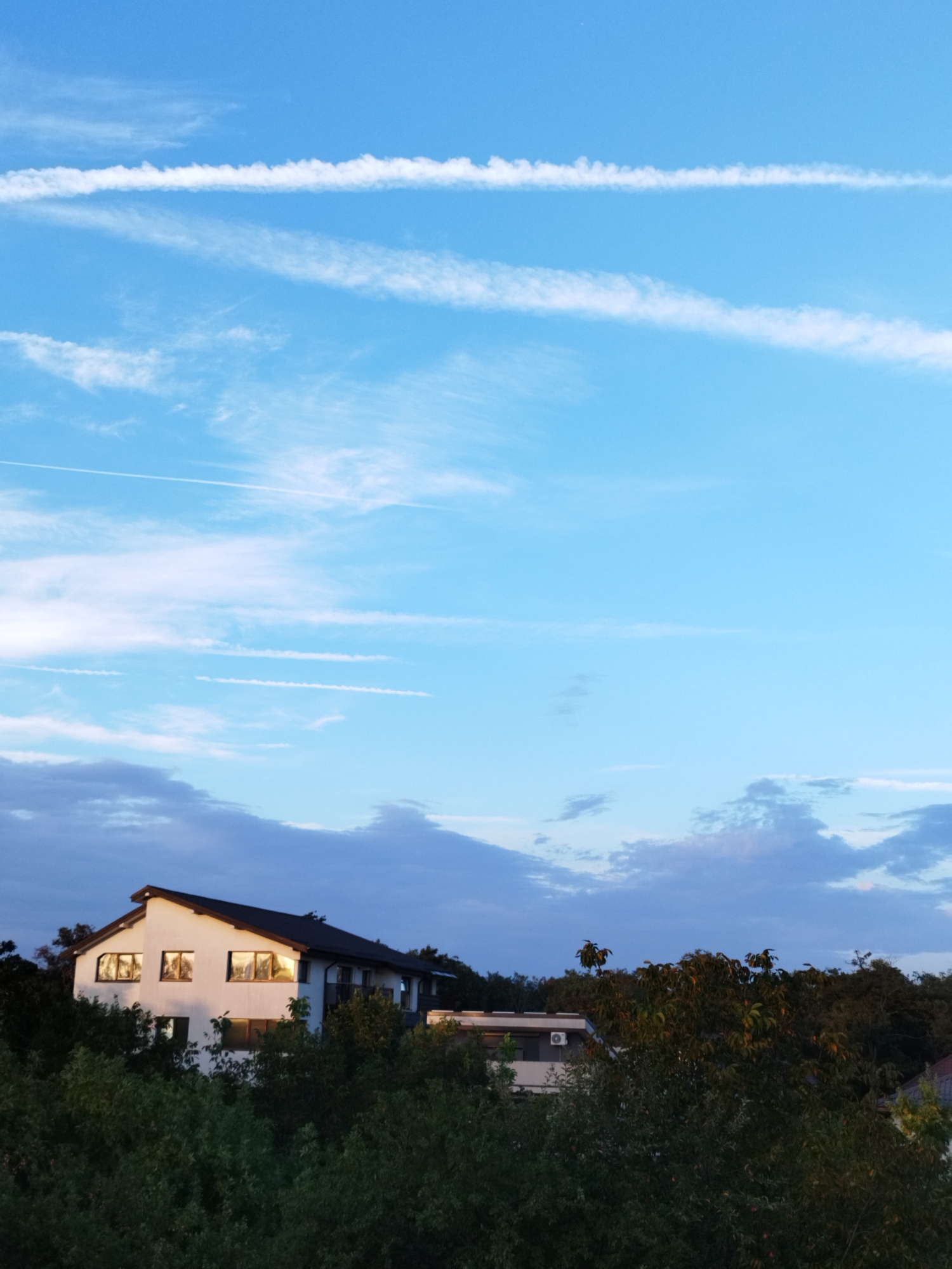 The image shows a white, two-story house with a dark roof, surrounded by lush green trees. The house has several windows that reflect the sky and what appears to be the setting sun, casting a warm, golden glow on the facade. The sky is a vibrant blue, with wispy white clouds and contrails from airplanes stretching across it. The setting appears to be a suburban or semi-rural area, possibly on the outskirts of Bucharest, Romania, given the time of day and the presence of both residential buildings and dense foliage. There are no people visible in the image.