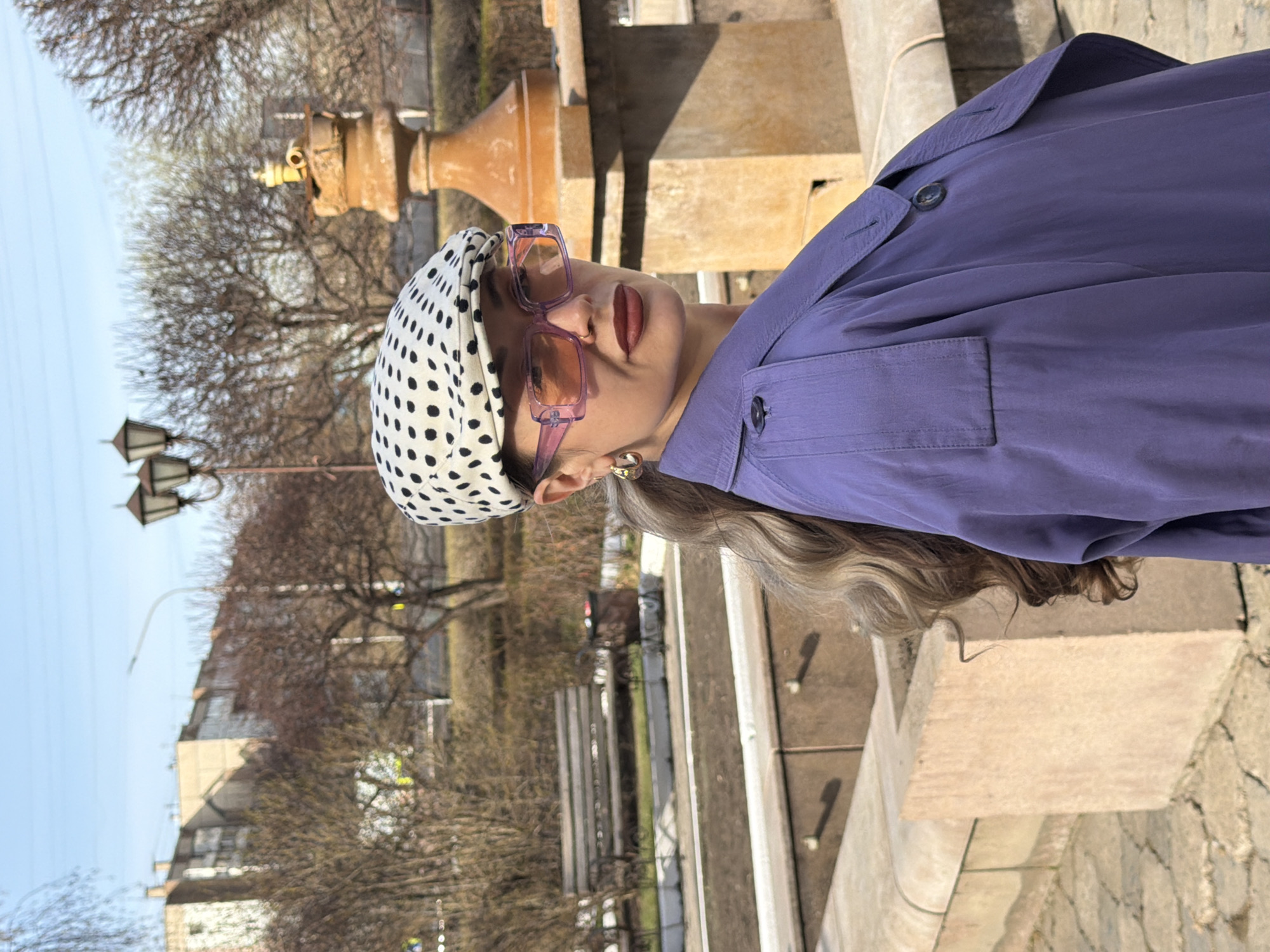 A medium shot, rotated 90 degrees clockwise, captures a person posing outdoors on a sunny day in what appears to be an urban park or square in Krasnoyarsk, Russia. The subject, likely female, is seen from the upper chest, wearing a stylish purple jacket or trench coat with visible buttons and a pocket detail. A white headscarf with black polka dots covers her head. She wears large, rectangular, translucent purple-framed sunglasses with tinted lenses, and dark red lipstick is visible beneath them. A gold-toned earring adorns her right ear, and long, wavy, light-toned hair falls behind her right shoulder.
The background features light-colored stone architectural elements, possibly part of a fountain or a balustrade. A distinctive, ornate metal object, resembling a vintage water spigot, is visible behind her head. Bare trees, indicating late winter or early spring, are prominent in the mid-ground, alongside several classic streetlights or lampposts. Further back, under a clear, light blue sky, are faint outlines of urban buildings. The ground appears to be paved with stone or concrete. The bright light suggests midday or early afternoon. No visible text is present in the image.