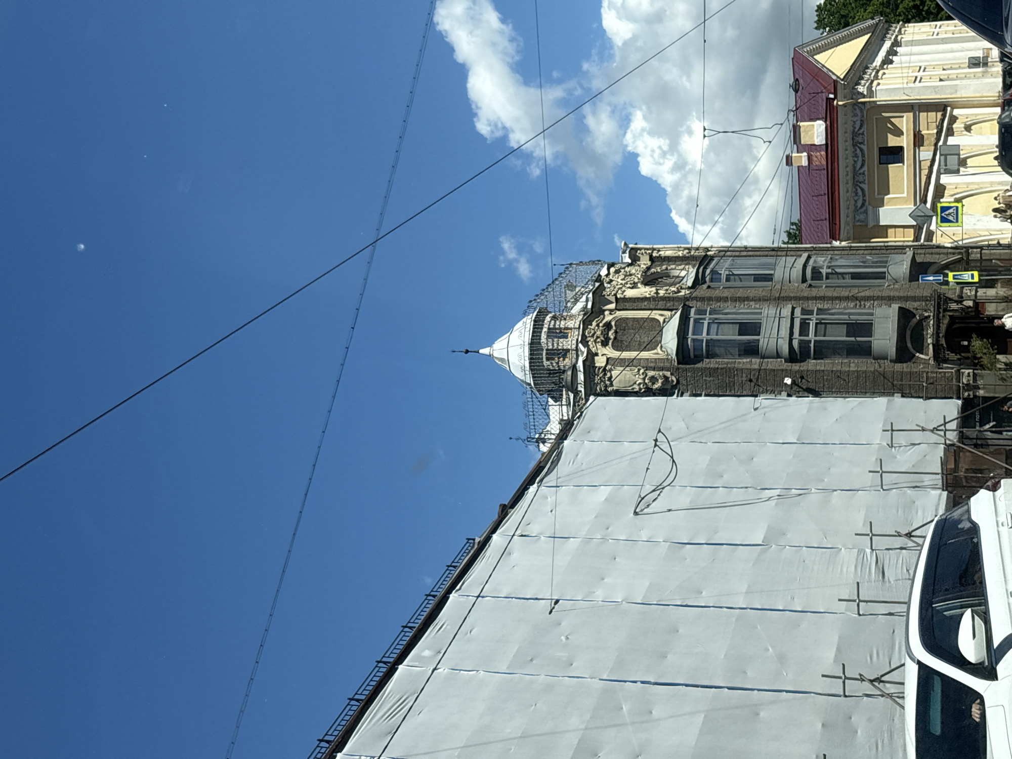 The image depicts an exterior view of buildings in Saint Petersburg, Russia, under a bright blue sky with scattered clouds. In the foreground, the side of a white car is visible. To the left, a large building is covered in white sheeting, likely for renovation or protection, supported by scaffolding. Adjacent to this covered building is a more ornate, older structure with intricate architectural details, including bay windows and decorative elements, topped with a white dome. To the right, a building with a red roof and yellow facade is visible, featuring columns and classical architectural elements. Power lines crisscross the sky above the scene. Several pedestrians are visible on the sidewalk near the yellow-roofed building. The overall impression is of an urban streetscape with historical architecture, some undergoing maintenance.
