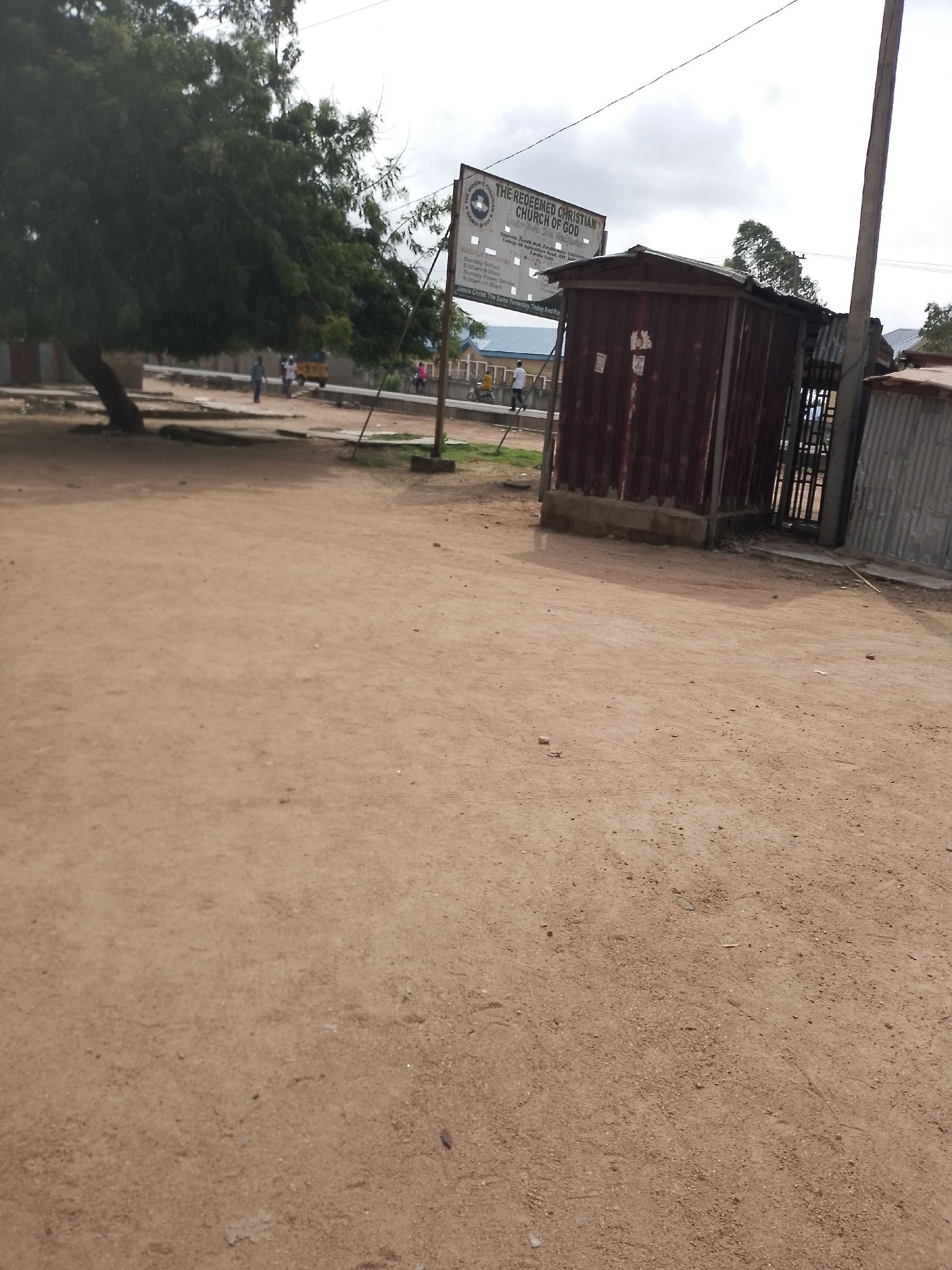 In an outdoor, dusty environment in Unknown, Unknown, a sign for "THE REDEEMED CHRISTIAN CHURCH OF GOD" is prominently displayed. The sign, attached to a pole, lists service times for Sunday school and other services. Adjacent to the sign is a small, weathered, reddish-brown metal structure with a pitched roof. To the right of this structure, a gate made of metal bars is visible, leading to an entrance. The ground is composed of loose, brown soil with scattered debris. In the background, a large tree with dense foliage stands to the left, and the blurred shapes of buildings and several people are visible. The sky is overcast.