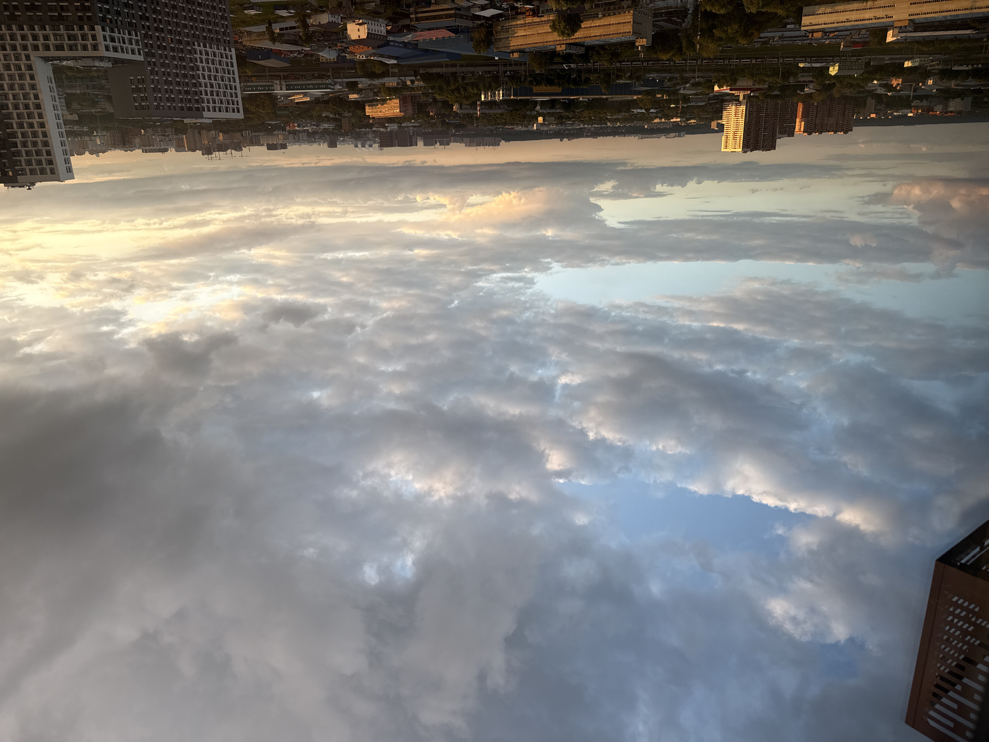 The image depicts an aerial view of a cityscape, likely taken from a high vantage point, possibly a tall building. The orientation of the image is rotated 90 degrees counter-clockwise. In the foreground, a vast expanse of clouds dominates the frame, with patches of blue sky visible between them. The clouds are varied in texture and density, with some appearing soft and wispy, while others are more layered and substantial. The lighting suggests it is either sunrise or sunset, as the clouds are illuminated by a warm, golden glow in some areas, contrasting with the cooler grays and blues in others.
In the background, a dense urban environment is visible. A prominent feature is a modern, high-rise residential building with a distinctive grid-like facade, seen on the far left. Further into the cityscape, numerous other buildings of various sizes and architectural styles can be discerned, including what appear to be industrial structures, some with long, rectangular roofs and rows of windows. The presence of trees interspersed amongst the buildings indicates a developed urban area with some green spaces. The horizon line shows a scattering of more distant buildings and infrastructure. The overall impression is one of a bustling city bathed in the dramatic light of either dawn or dusk. The weather appears partly cloudy, with the clouds creating dynamic shadows and highlights across the sky and the urban landscape. There is no visible text in the image.