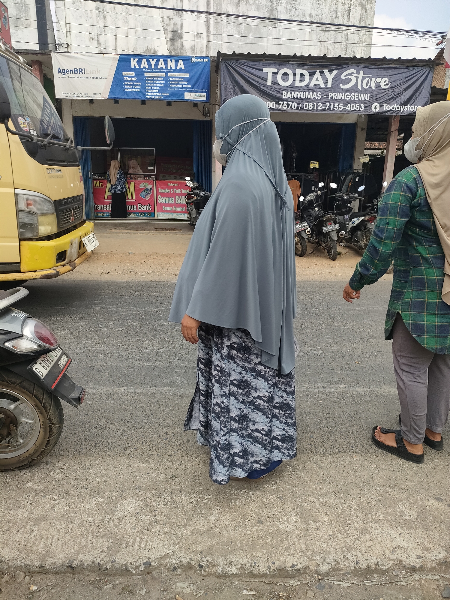 In Sukoyoso, Indonesia, a street scene unfolds in front of commercial establishments. A yellow truck is parked on the left, partially obscuring a storefront with signage that reads "AGENBRI Link" and "KAYANA." The latter displays services like "T-BANK," "BAYAR LISTRIK," "BAYAR TELKOM," and "CEK SALDO." Further down the street, a large banner advertises "TODAY Store," with contact information and a Facebook handle. Two women, both wearing hijabs and face masks, are visible. One woman, in the foreground and facing away from the camera, wears a grey hijab and a long, patterned skirt. The second woman, on the right and facing towards the store, is dressed in a green and black plaid shirt and grey pants. Several motorcycles are parked in front of the "TODAY Store." The street is paved with asphalt and shows signs of wear. A motorcycle license plate is partially visible on the left side of the image. The overall environment suggests a bustling commercial area.
