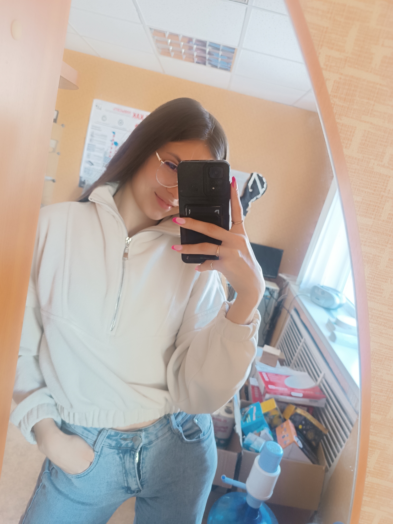 A young woman is taking a selfie in a mirror indoors. She is wearing glasses, a light-colored pullover with a short zipper, and blue jeans. Her nails are painted with a light pink French manicure. She is holding a dark-colored smartphone in her right hand. The background is cluttered with various items, including boxes and a water dispenser, suggesting a storage or work area. A window is visible to the right, and a poster hangs on the wall behind her. The location is an unknown interior in the city of Unknown, Unknown.