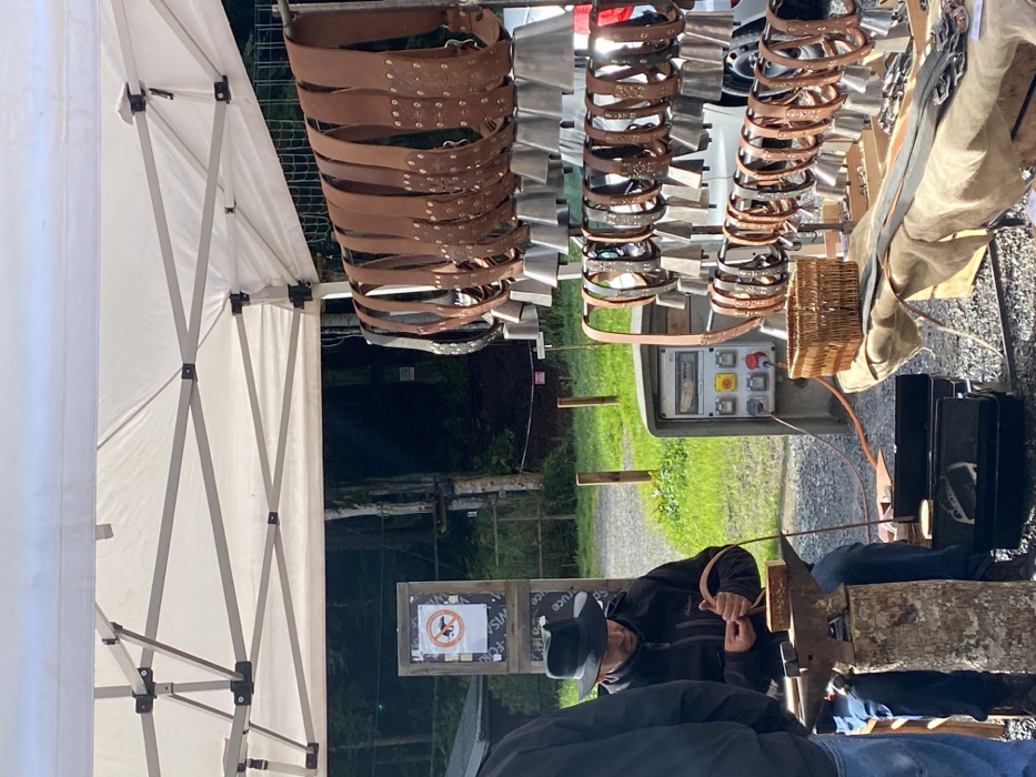 The image captures an outdoor market stall or craft demonstration situated in Abondance, France. Under a white fabric canopy supported by a metal framework, a male individual wearing a dark fedora-style hat, dark zippered jacket, and blue jeans is seated on a stool at the left. He is actively engaged in crafting, manipulating a piece of brown leather with both hands over a small, dark-colored anvil that rests on top of a vertical wooden log acting as a stand. Another individual's left torso and arm are partially visible on the extreme left, facing towards the craftsman.
To the right of the craftsman, a multi-tiered display rack holds numerous brown leather belts, each adorned with multiple metal bells of varying sizes, commonly known as cowbells. These belts are arranged horizontally across several metal rods. Below this primary display, additional brown leather straps and metal buckles are laid out on a light-colored fabric covering a lower surface. A small, light-brown wicker basket is positioned on this surface. Behind the display, an industrial-style grey electrical control panel with a top-mounted gauge, red circular buttons, and yellow toggle switches is visible, with an orange power cord extending from it towards the left.
The background reveals an outdoor setting: a light-colored gravel path extends from left to right, bordered by green grass and sparse wooden posts. Beyond the path, a dense, dark-green wooded area with tall trees forms the backdrop. A square wooden sign depicting a red circle with a diagonal line over a dog icon, signifying "no dogs allowed," is mounted on a wooden post near the path. The scene is illuminated by natural light, indicating daytime.