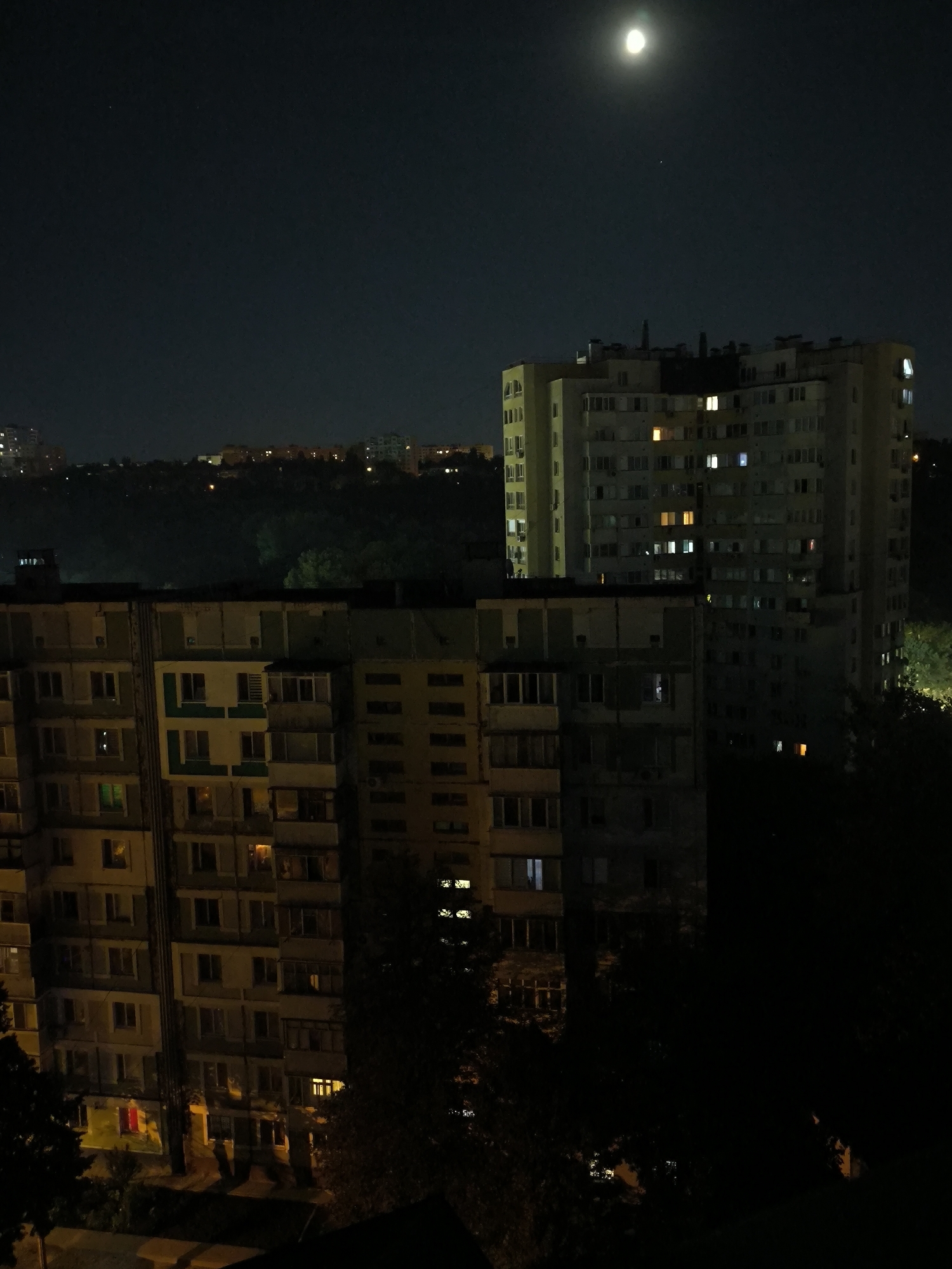 The image shows a nighttime view of apartment buildings in Chișinău, Moldova. The moon is visible in the dark sky, illuminating the scene. The buildings are multi-story structures with many windows, some of which are lit, suggesting that people are inside. The architecture appears to be typical of Soviet-era apartment blocks. Trees and other vegetation are visible in the foreground and between the buildings, and a hilly landscape with more distant buildings can be seen in the background. The overall impression is of a residential area at night, with the moon casting a soft glow.