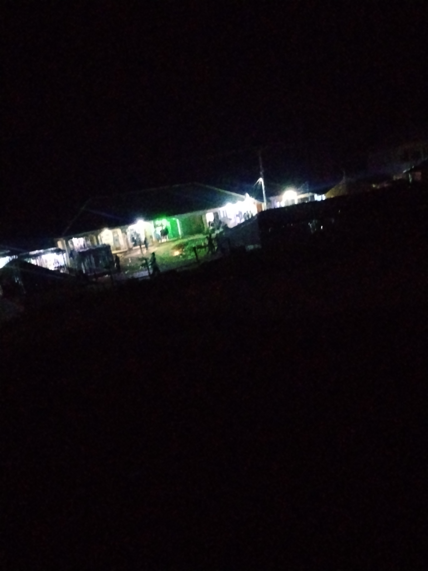 The image depicts a nighttime scene in what appears to be a market or gathering area in the city of Durumi, Nigeria. Several buildings are illuminated with artificial lights, suggesting commercial activity or residential structures. Some people are visible, standing or walking, though their features are indistinct due to the low light conditions. The ground appears to be unpaved and possibly wet in some areas, indicating it might be an outdoor market or a less developed urban space. Several vehicles, possibly small trucks or stalls, are also present, further contributing to the impression of a lively, albeit dimly lit, evening gathering. The overall ambiance is one of a typical Nigerian community coming to life after dark.
