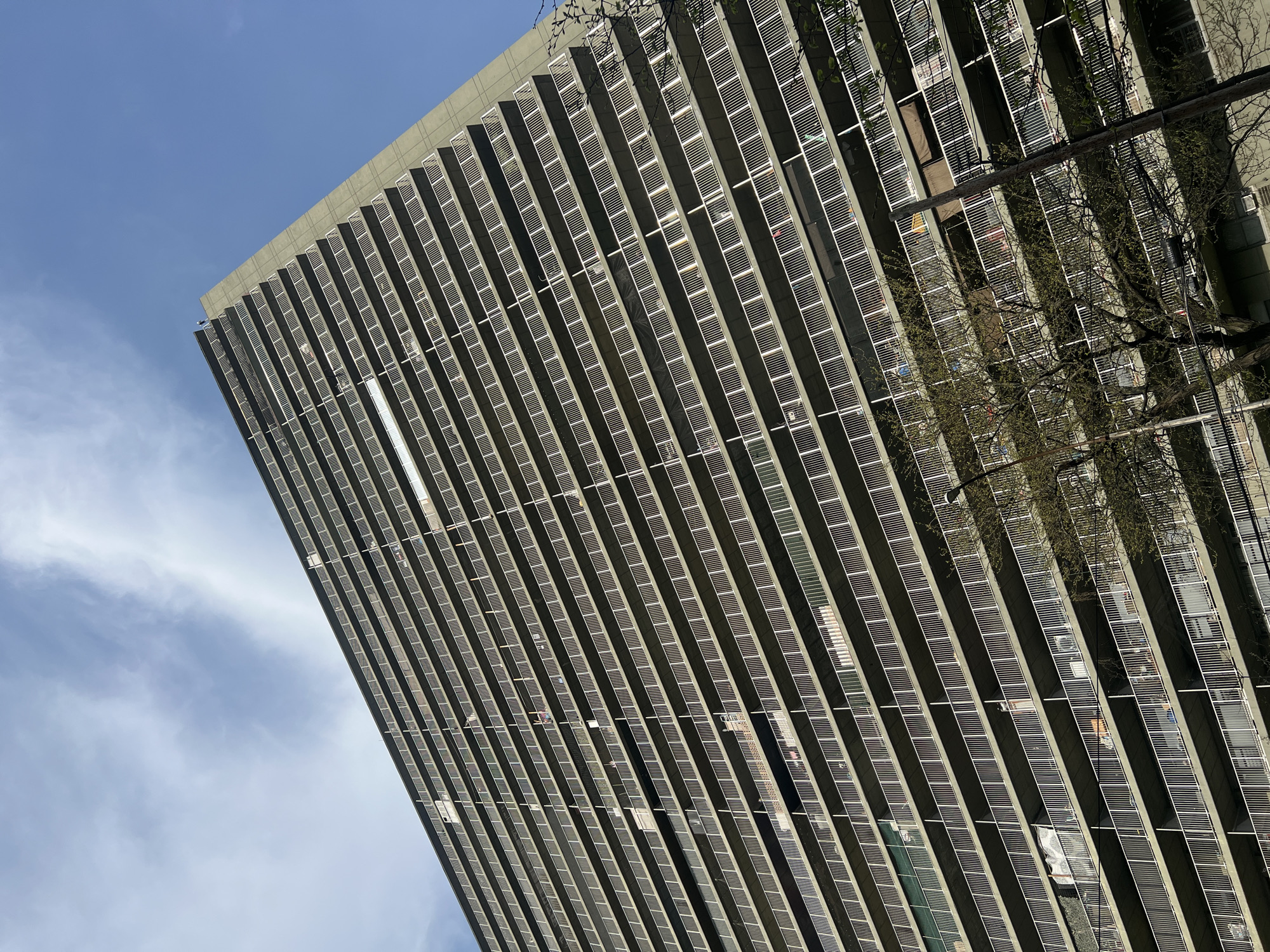 The image shows a tall building, likely an apartment complex, viewed from a low angle. The building has a repetitive pattern of windows with white, horizontal shades or blinds. A few windows are open. A tree branch is visible in the top right corner. The sky is blue with some clouds. The location is likely New York City, United States of America.