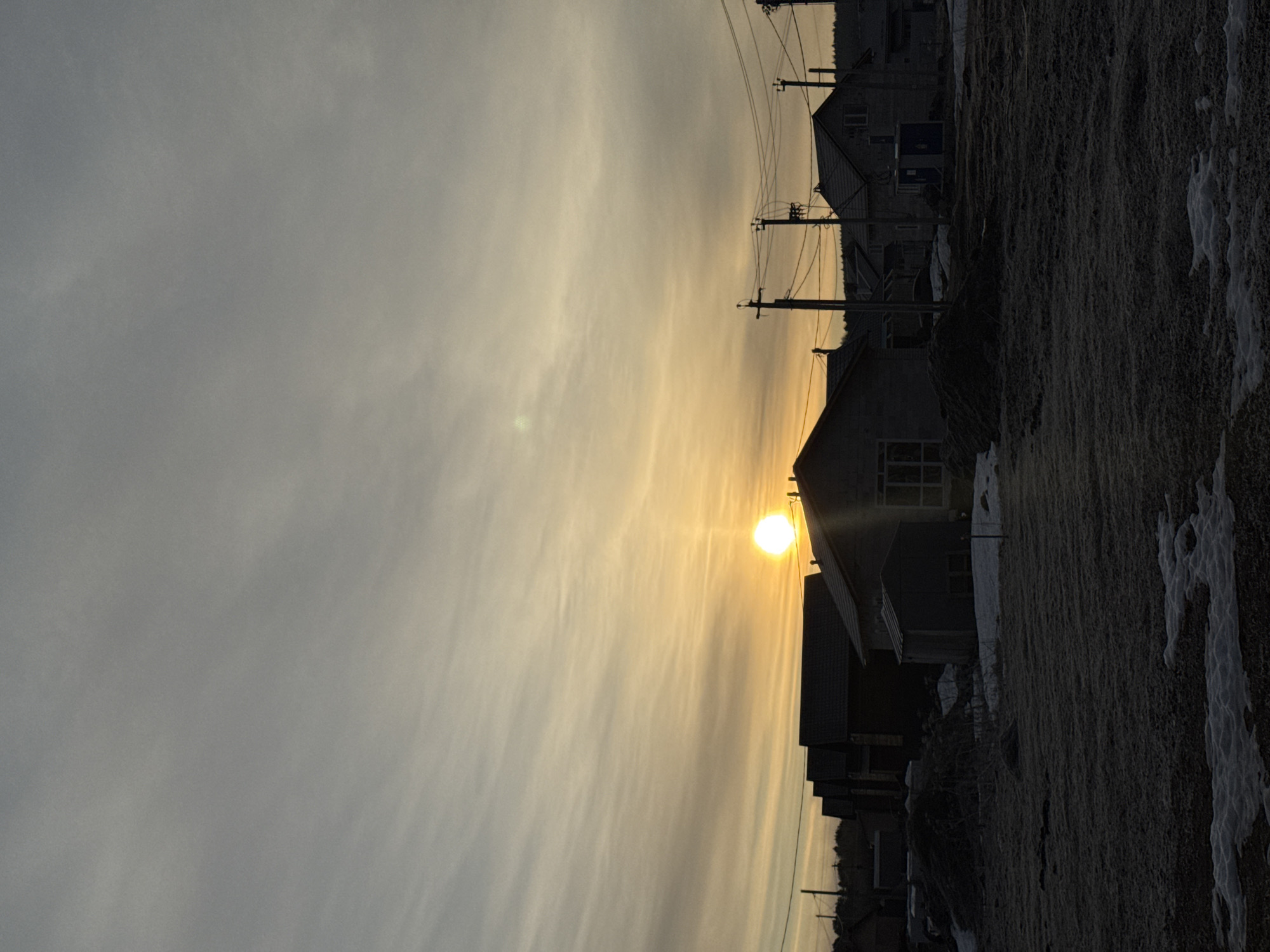 The image shows a rural setting in Nestiukovo, Russia at dawn or dusk. The sun is low in the sky, casting a warm, yellow glow. Several houses are visible, silhouetted against the bright light. Power lines stretch across the frame, connecting the buildings. The ground appears uneven and partially covered with patches of snow. There are no people visible.