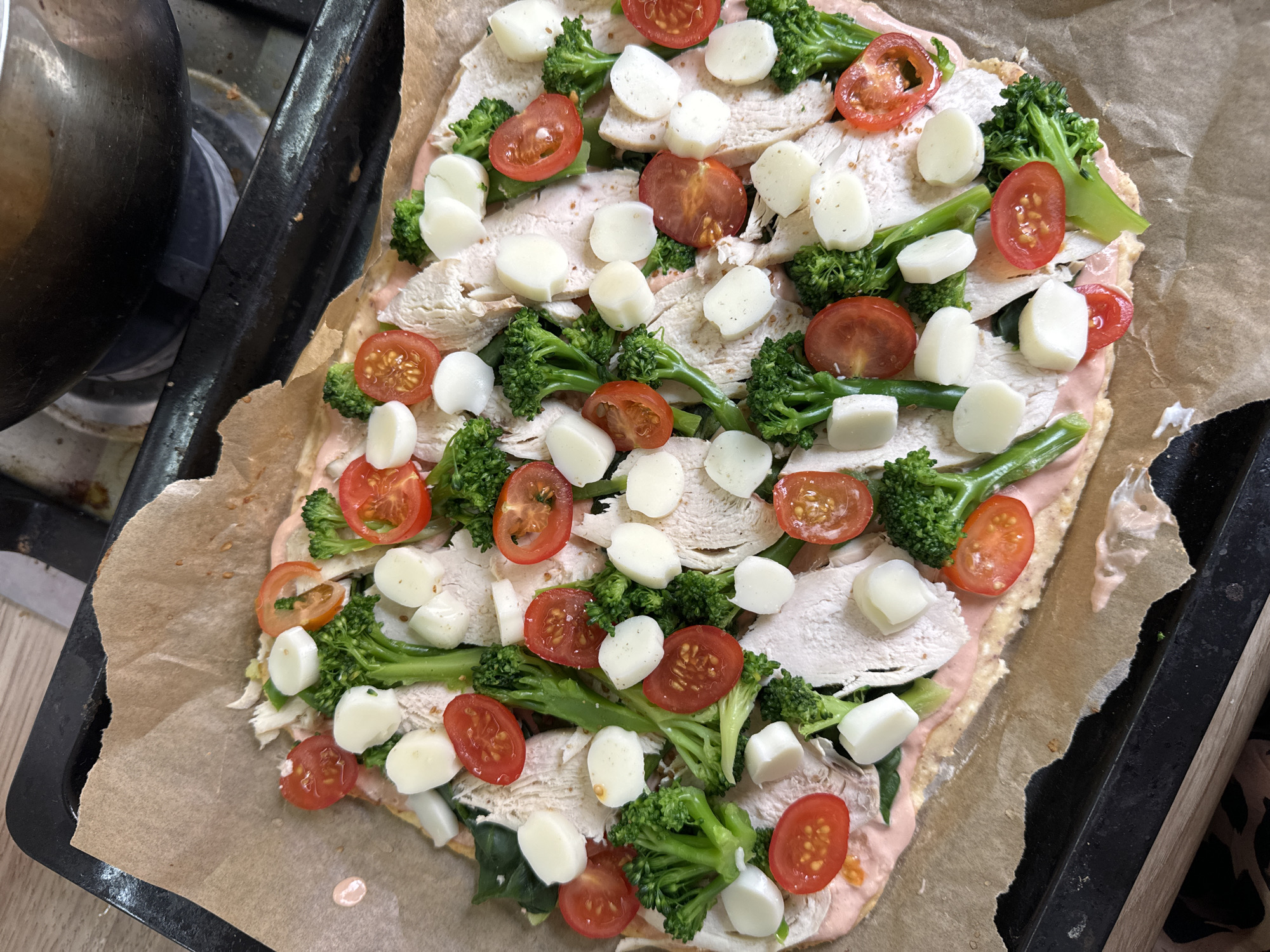 The image shows a pizza on a baking sheet lined with parchment paper. The pizza is topped with what appears to be a light pink sauce, sliced cooked chicken, broccoli florets, halved cherry tomatoes, and small, round pieces of what appears to be cheese. The baking sheet is sitting on a dark stovetop next to a stovetop burner. The environment appears to be a home kitchen. There is no indication of Krasnoyarsk, Russia, in the image.