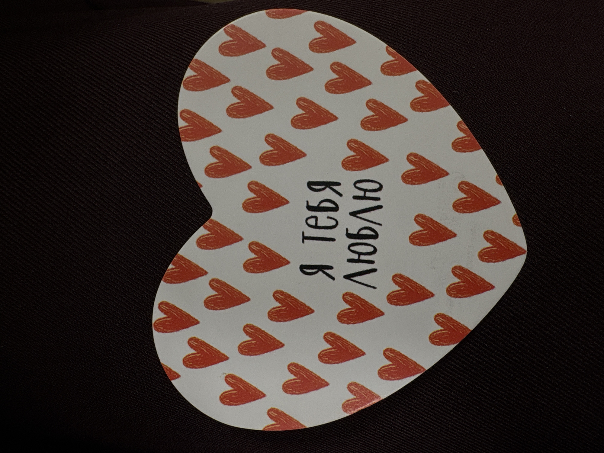 A heart-shaped card with a pattern of small, red, hand-drawn hearts is placed on a dark, textured fabric. The card features a message in black Cyrillic text in the center, which reads "Я тебя люблю" (Ya tebya lyublyu), meaning "I love you." The card is not located in a specific place in Saint Petersburg, Russia; it is a standalone object.