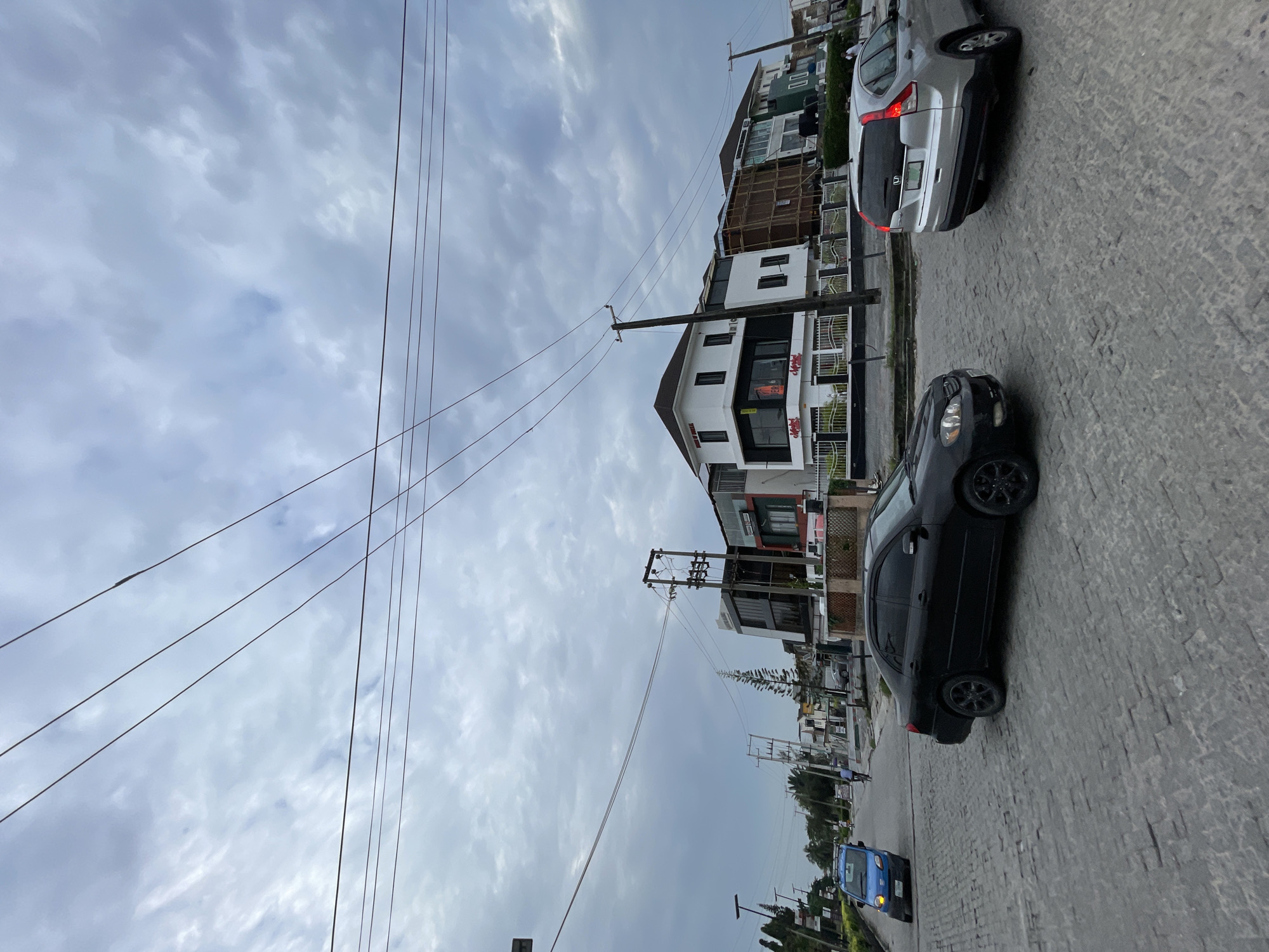 The image depicts a street scene in Moba, Nigeria, characterized by a cloudy sky and a paved road. Several modern buildings line the street, featuring white and dark facades with large windows. Power lines crisscross the sky above the street. Two cars are visible on the road: a black sedan in the foreground and a silver SUV in the mid-ground. A blue van is also present on the left side of the street. In the background, more buildings and trees can be seen, along with utility poles and streetlights. The overall impression is one of an urban environment under overcast conditions.