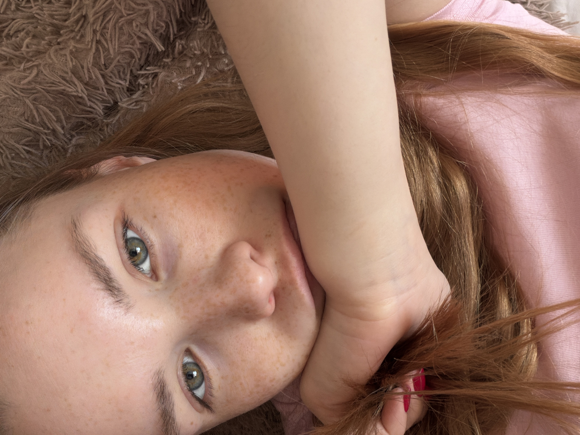 A close-up image features a young woman with fair skin and freckles, lying down. Her hair is reddish-brown, and she is wearing a pink top. Her left hand is holding a portion of her hair near her face. The background consists of a soft, brown, fluffy material resembling a rug or blanket. The location in the city of Berezniki, Russia is not discernible from the image.