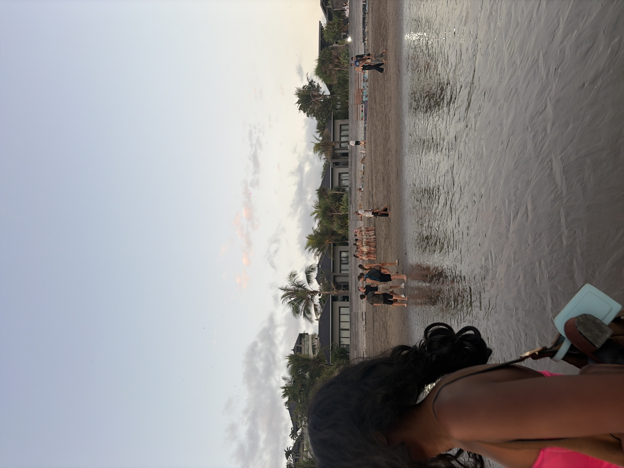 In North Kuta, Indonesia, a group of individuals is gathered on a beach near the ocean. The sky is a pale blue with scattered clouds, and the sun is low, casting a warm light. The sand is wet and reflects the sky and surrounding buildings. In the foreground, a person with dark, curly hair is visible from the side, wearing a bright pink top and carrying a bag. Further down the beach, a group of people stands near the water, some wearing swimwear and others casual clothing. Behind them, a row of modern buildings with dark roofs is visible, surrounded by lush palm trees and tropical vegetation. The overall scene suggests a relaxed, tropical atmosphere, possibly during sunset or early morning.