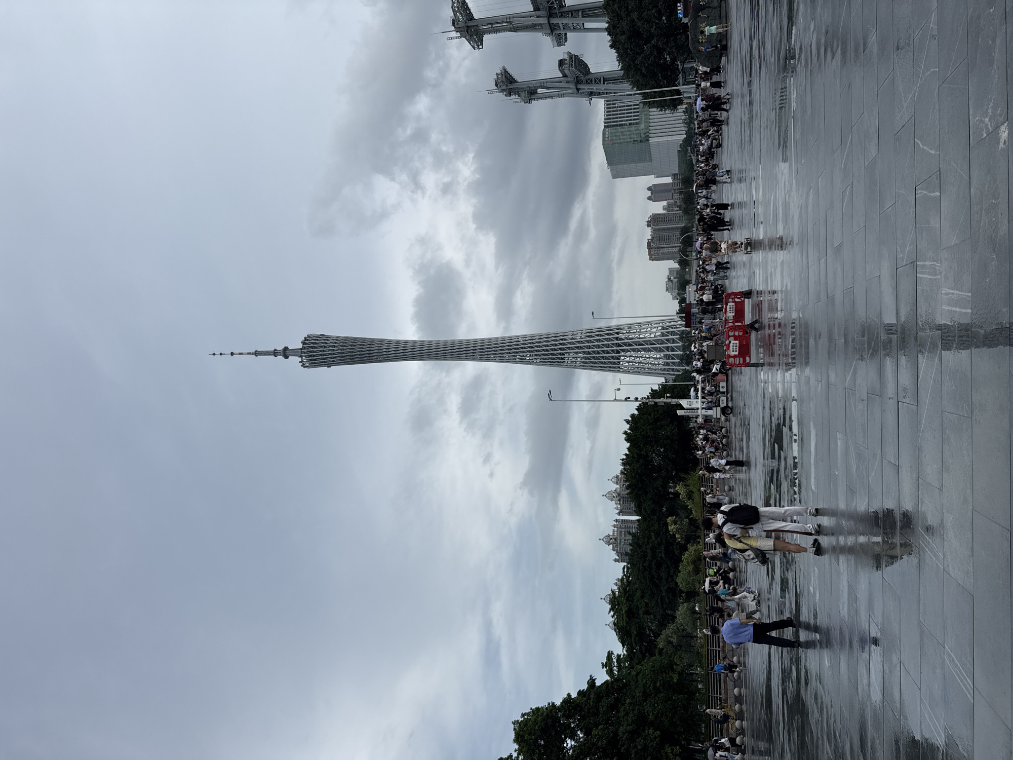 This is a wide-angle, eye-level shot of the Canton Tower area in Tianhe District, China, on a cloudy day. The Canton Tower, a distinctive lattice-like structure, dominates the center of the frame, reaching towards the overcast sky. In the foreground, a wide, wet, stone plaza reflects the ambient light and the figures moving across it. Several people are visible scattered across the plaza, some walking individually, others in pairs or small groups. Two figures in the immediate foreground are embracing.
To the right of the Canton Tower, two large, industrial-looking structures with exposed frameworks stand prominently. In the background, a dense urban landscape unfolds, with various buildings of different heights and architectural styles, including a modern glass skyscraper. To the left of the tower, a line of lush green trees separates the plaza from the distant cityscape. The ground is wet, indicating recent rain or ongoing drizzle, and a few people are holding umbrellas, suggesting damp conditions. The overall atmosphere is subdued due to the cloudy weather. A few wheeled vehicles, possibly for rent or for transportation, are also visible in the middle ground.