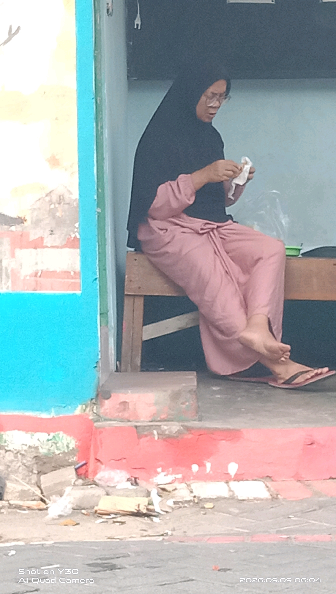 A woman wearing a black hijab and a pink garment sits on a wooden bench on a raised platform outside a building. She is holding a white tissue in her hands and appears to be wiping something. She is wearing glasses and flip-flops. The background shows a blue wall on the left and a light blue wall on the right. In front of the platform is a street with some debris on the ground. The scene appears to be located in a public space, possibly a market or a street-side shop. The timestamp on the image indicates it was taken on September 9, 2026, at 06:04. This location is identified as Klangonan, Indonesia.