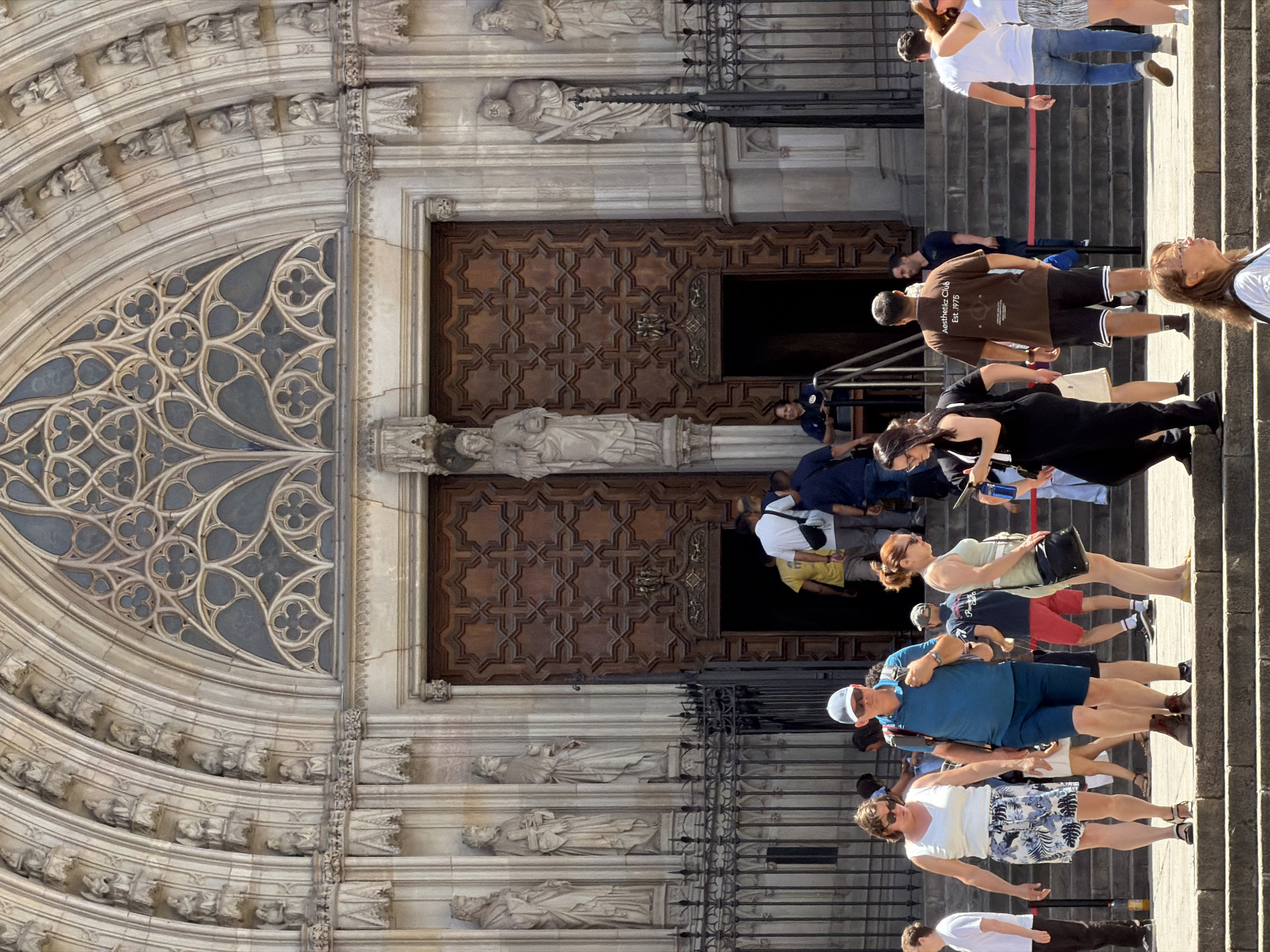 The image shows the entrance to the Barcelona Cathedral, a Gothic cathedral located in the city of Barcelona, Spain. The cathedral's facade features intricate stone carvings, statues, and a large rose window. In the foreground, a group of tourists is seen walking up the stairs towards the entrance. Some are dressed casually, while others appear to be dressed more formally. The tourists are interacting with each other, with some pointing and talking, while others are taking pictures. The atmosphere appears to be lively and busy, with people enjoying the sights and sounds of the city. The weather is sunny, and the sky is clear.