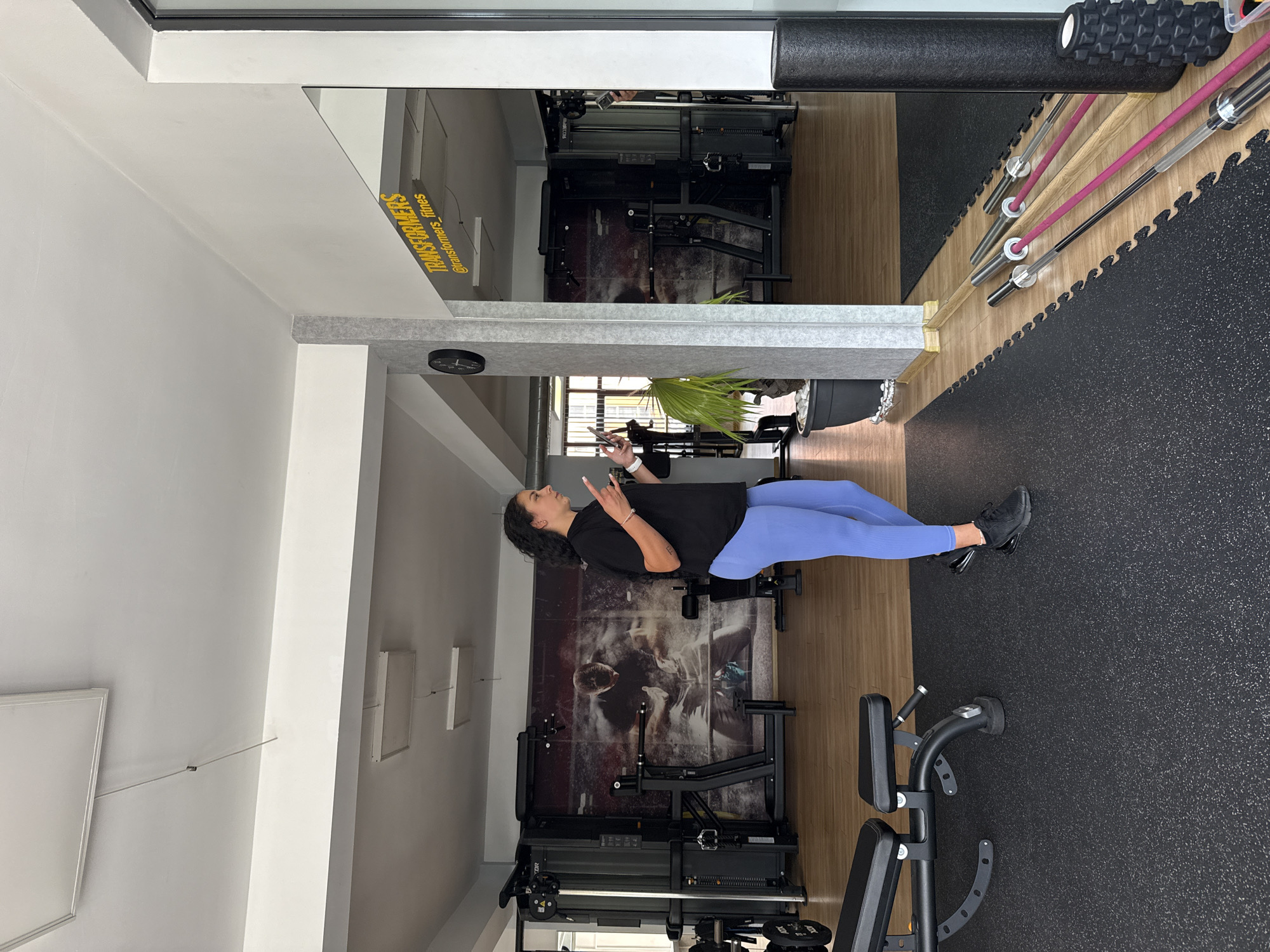The image depicts the interior of a gym, likely located in Gabrovo, Bulgaria, as indicated by the presence of "TRANSFORMERS_fitnes" signage. A woman with dark curly hair, wearing a black t-shirt and blue leggings, is standing and gesturing with her hands. She appears to be talking or explaining something, holding a phone in one hand and a small object in the other.
The gym features various exercise equipment, including weight machines, a weight bench, dumbbells, and weightlifting bars. A large mural of a weightlifter is visible on the back wall. The floor is covered with black rubber mats and lighter wood-toned flooring in different sections. A large potted plant is situated near a pillar. The environment is well-lit with overhead fluorescent lighting. A mirror reflects part of the gym, including more equipment and the signage. The overall scene suggests a functional and active fitness facility.
