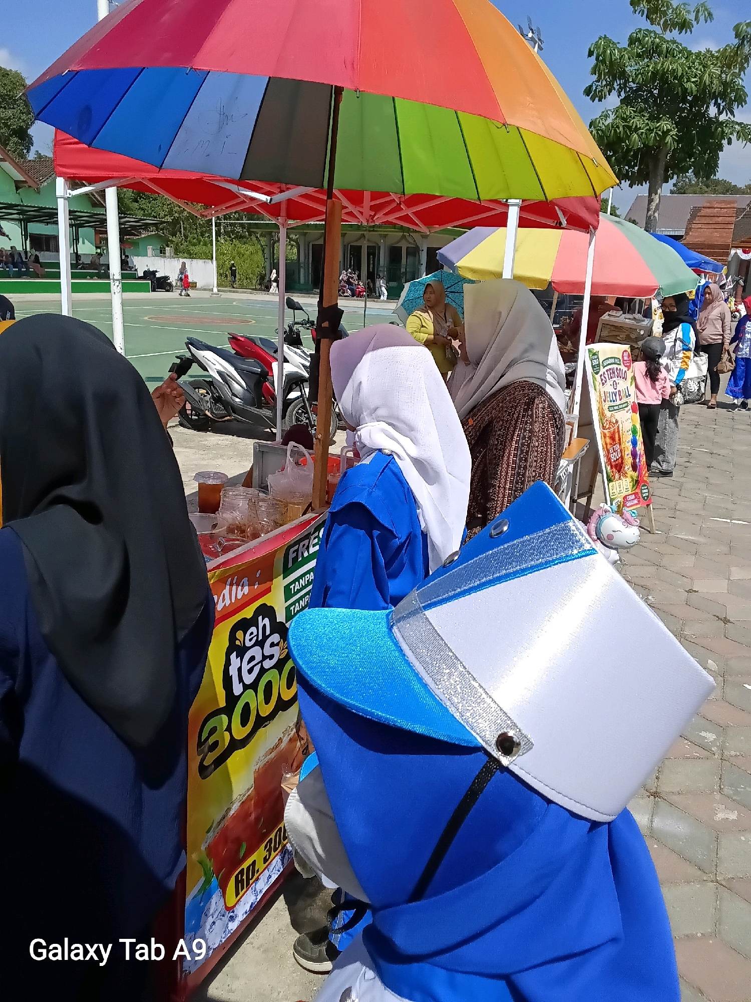 A street scene unfolds in the city of Ngadirenggo, Indonesia, under a clear sky. The foreground is dominated by the back of two individuals wearing blue uniforms, one with a white cap adorned with silver trim. They appear to be standing near a food stall that features a colorful banner advertising "Es Teh Solo" for Rp. 3000. Behind them, a variety of umbrellas are set up, casting colorful shades. In the middle ground, a basketball court is visible, with a few people sitting and standing around. Several motorcycles are parked near the court. Further back, buildings with green roofs and trees line the area, suggesting a public or recreational space. A few more people are seen in the background, some seemingly engaged in activities near the stalls and buildings. The overall atmosphere suggests a daytime outdoor gathering or event.