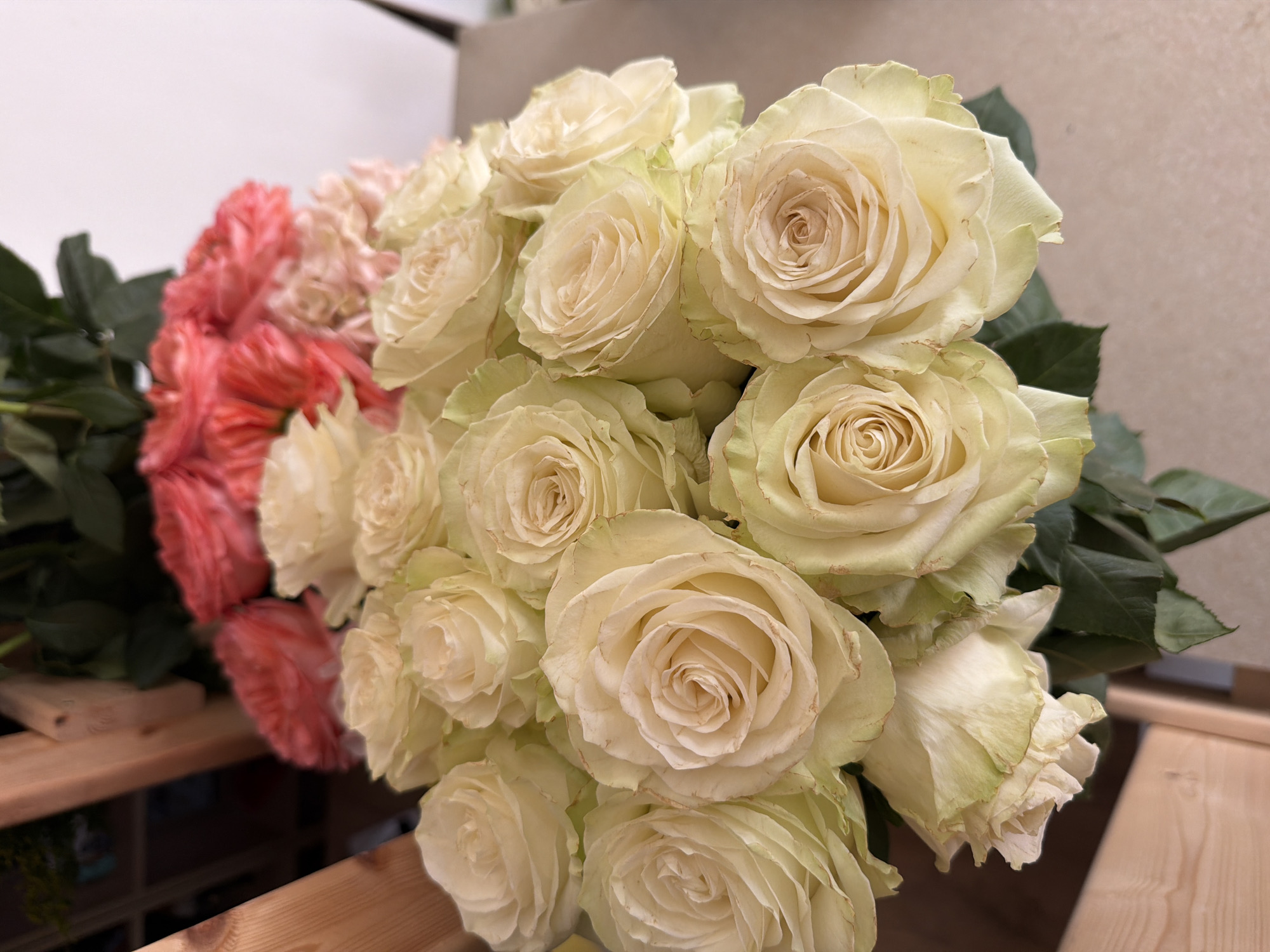 A bouquet of roses, primarily in shades of cream and pale yellow, is displayed in the foreground. Behind these, a cluster of coral and light pink roses is visible, creating a colorful contrast. The arrangement is set against a neutral background, with hints of wooden elements and greenery on the left side, suggesting an indoor floral setting, possibly a flower shop or a home. The image does not provide specific details about its location in Khimki, Russia.