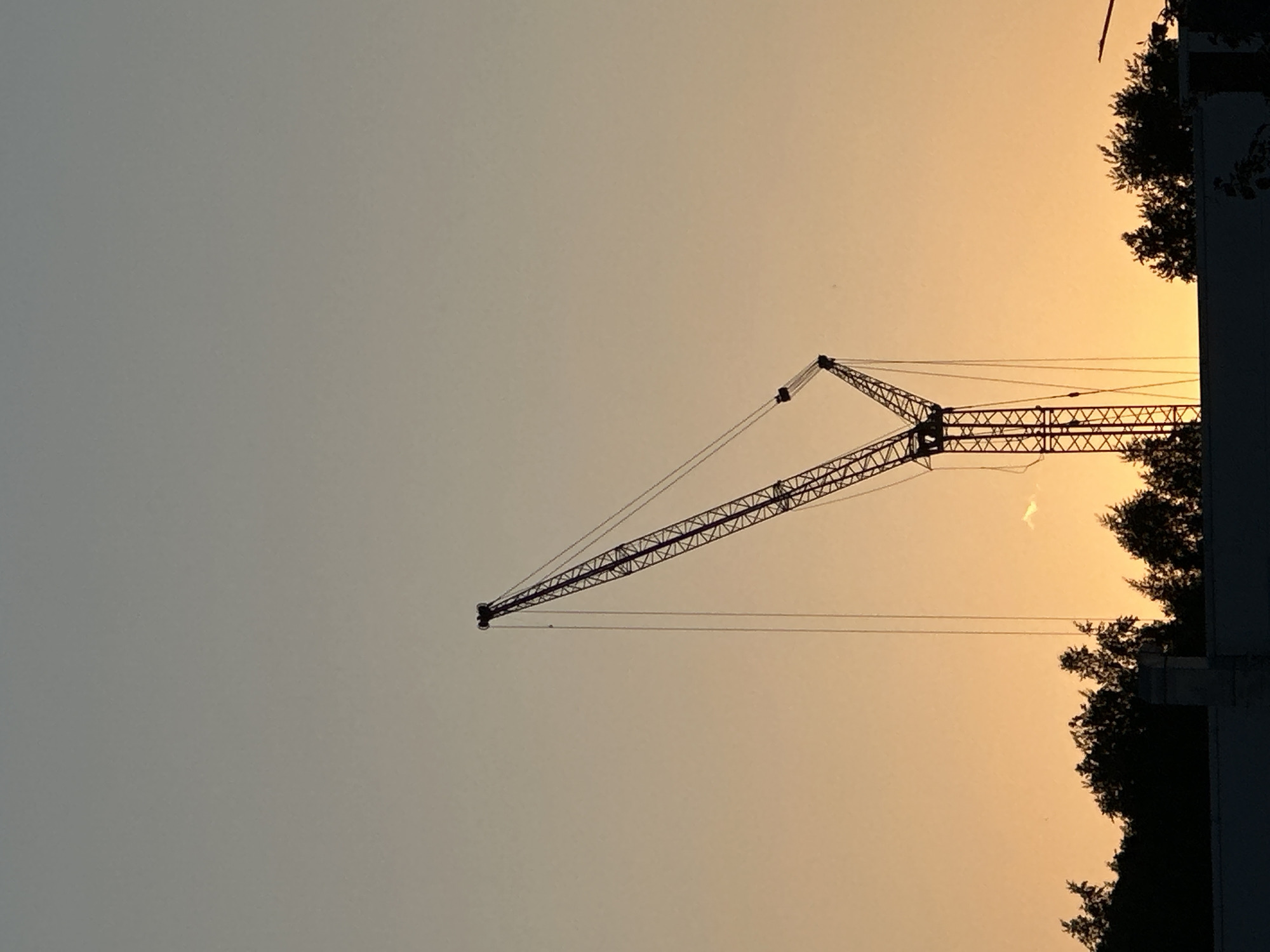 The image shows a construction crane silhouetted against a gradient sky, ranging from light gray to orange, likely at sunset or sunrise. The crane is positioned off-center to the left of the frame. To the right, there are trees and a building outline. The location is unidentified.