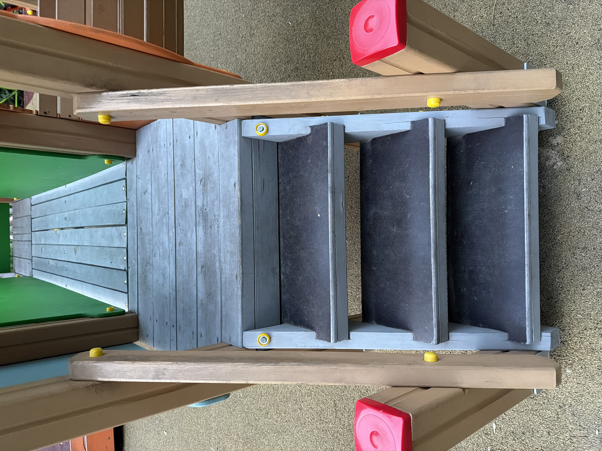 A close-up, eye-level shot shows a set of three steps leading up to a wooden platform of a playground in Shymkent, Kazakhstan. The steps are painted light gray with dark gray treads. Yellow plastic caps are visible on the sides of the steps and on the adjacent wooden supports. On either side of the steps, at the base, are bright red, square-shaped plastic elements, each with a circular indentation in the center. The ground surface appears to be a light brown, textured material, likely rubberized safety surfacing common in playgrounds. The background reveals more of the playground structure, including wooden beams, a green panel, and a slatted wooden walkway extending further up. The lighting suggests an overcast day. No people are present in the frame.