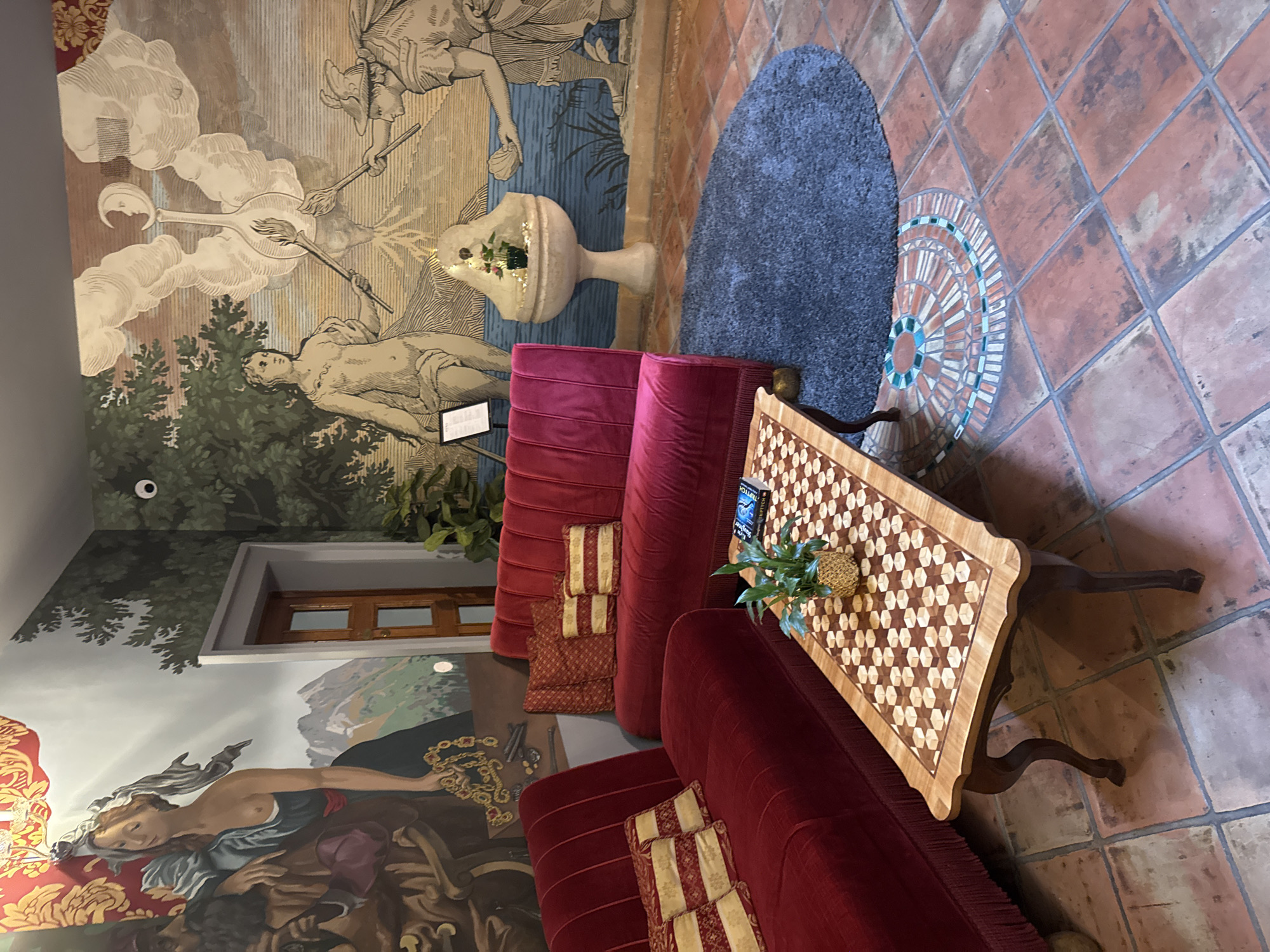 The image depicts an interior scene with opulent decor. In the foreground, a red velvet sofa with tufted upholstery and fringe accents is arranged in an L-shape. Several decorative pillows with a gold and red pattern are placed on the sofa. In front of the sofa is a wooden coffee table with an inlaid checkerboard pattern on its surface. A small potted plant sits in the center of the table, next to a book.
The background features a large, detailed mural covering the walls, depicting a mythological or allegorical scene. The mural includes figures of people, animals, and celestial bodies, with a flowing landscape and water elements. To the right of the mural, a decorative pedestal sink with a small plant and lights is visible.
A plush, dark gray circular rug is placed on the terracotta tile floor, partially in front of the sofa. The floor itself is tiled with large, square terracotta tiles, with some decorative mosaic patterns near the rug.
The lighting appears to be a mix of ambient and possibly some artificial lighting, creating a warm and inviting atmosphere. The overall impression is one of a luxurious and art-filled space. The specific location in Prague, Czechia is not discernible from the visual information provided.