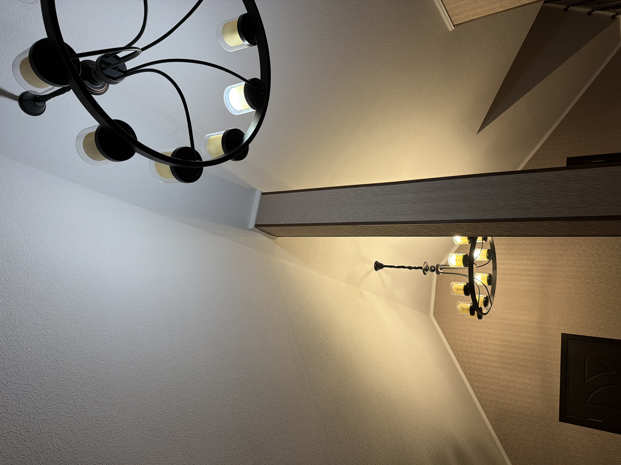 The image shows the interior of a building with a textured ceiling and sloped walls. Two chandeliers are visible. One is a large, black, multi-armed chandelier with candle-shaped bulbs, hanging from the upper right of the frame. Another, smaller chandelier with a similar design is visible on the left side of the frame. The walls are covered in a textured wallpaper in a neutral tone, and a dark wooden door is visible in the lower left corner. A tall, vertical beam with a wood-grain texture is positioned in the center of the image, dividing the space. The lighting suggests it is either evening or the space is designed for ambient illumination. The scene is static and depicts an interior architectural space.