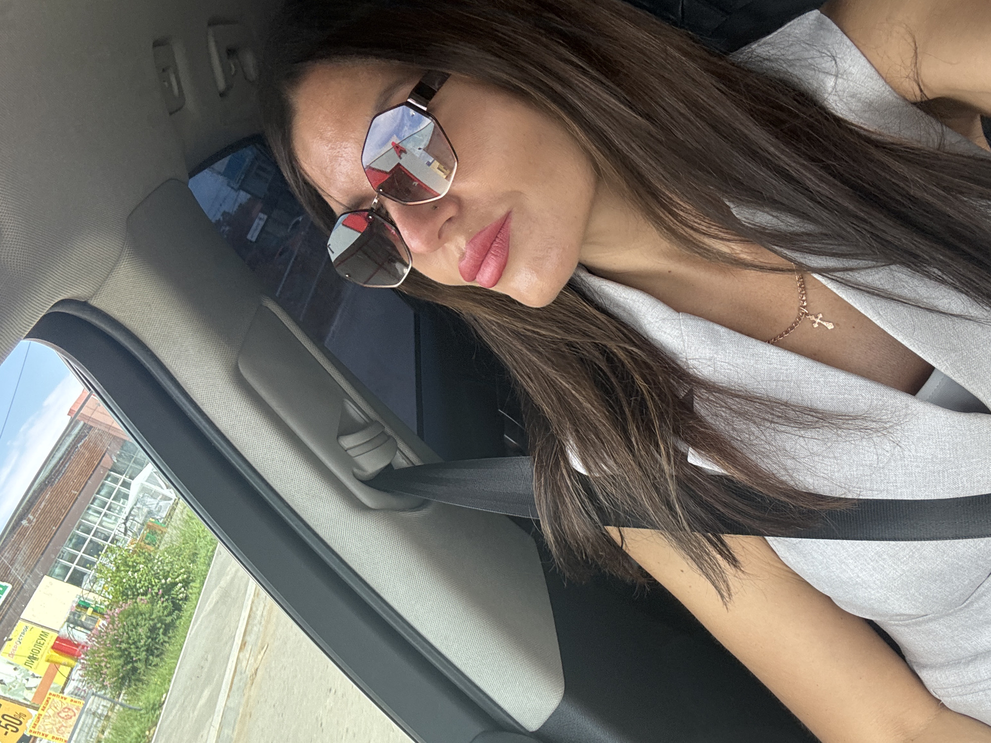 A woman wearing sunglasses and a light-colored vest is sitting in the passenger seat of a car, looking to her left. She has long, brown hair and is wearing a necklace with a cross pendant. The interior of the car, including the seatbelt and headliner, is visible. Through the car window, buildings, vegetation, and a clear sky can be seen. The exterior scene appears to be a commercial or industrial area, possibly in Chaikovsky, Russia, as indicated by the signage visible on one of the buildings which includes Russian text, for instance, "ДОБРОСТРОЙ" (Dobrostroy).
