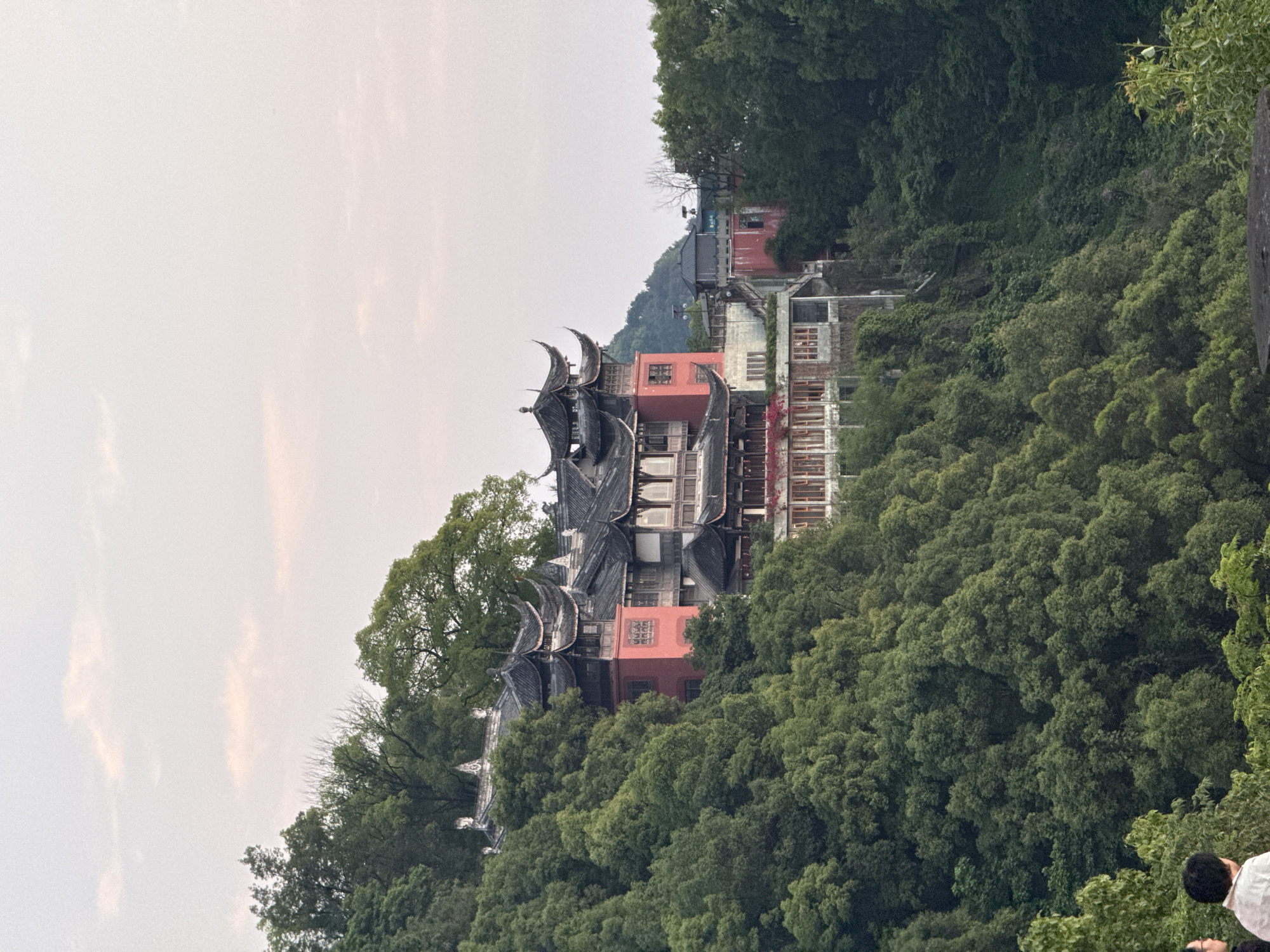 The image depicts a multi-tiered traditional Chinese building, likely a temple or historical structure, nestled amidst lush green foliage. The building features dark, intricately designed roofs with curved eaves and ornamental details. Red and white painted walls are visible between the levels. The sky is overcast. The structure is located in Nan'an District, China. A person is seen in the lower right corner.