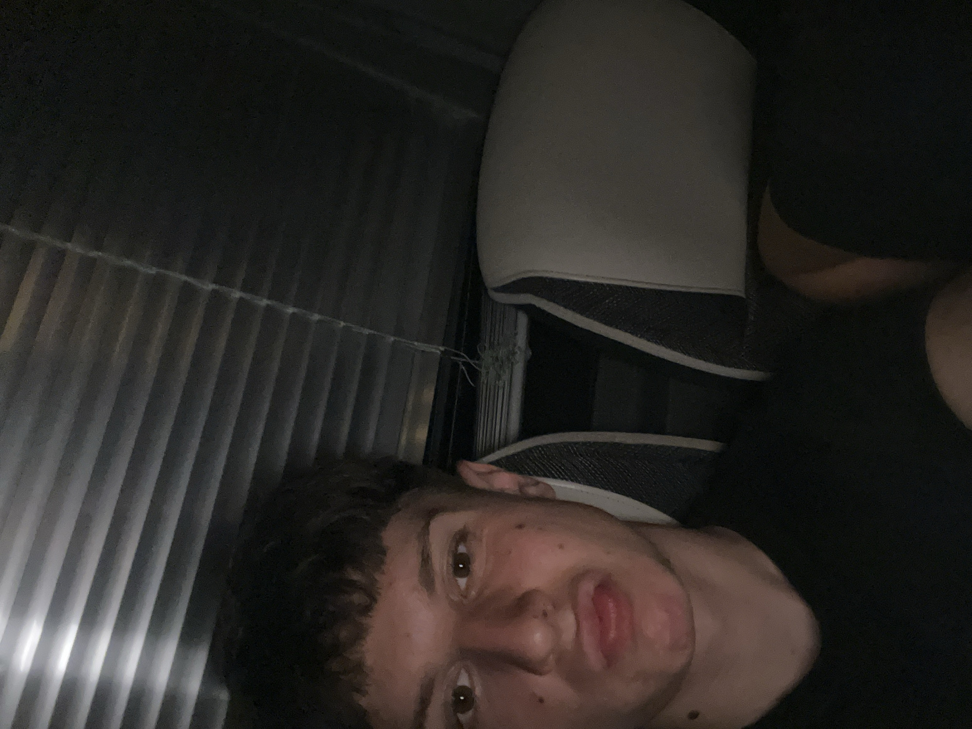 The image depicts a person with dark curly hair, light skin, and brown eyes lying down. The person's head is tilted to the side. Their mouth is slightly open. The background includes a corrugated metal wall, and what appears to be the back of a car seat. The environment is dimly lit. It is not possible to determine the location within Milevo, Bulgaria, or any specific actions or interactions from the image alone.