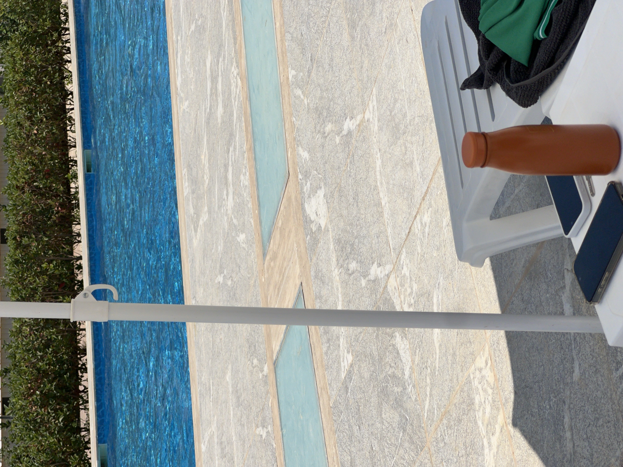 In Aksu, Turkey, a white umbrella pole stands in the foreground, casting a shadow on the tiled poolside. To the right, a white lounge chair is partially visible, with a dark towel and a green fabric draped over it. A brown water bottle and two smartphones rest on the seat of the chair. In the background, a clear blue swimming pool is bordered by lush green bushes.