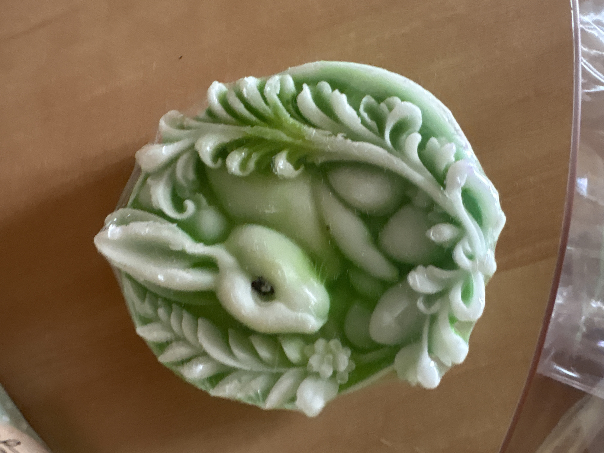 This is a close-up, overhead shot of a green and white decorative soap bar shaped like a rabbit surrounded by foliage. The soap is sitting on a light brown surface, possibly a table. The rabbit is in a reclined position, facing left with its ears up. The foliage appears to be made of leaves and small flowers, creating a circular frame around the rabbit. The lighting is somewhat dim, casting a soft glow on the soap. The background is out of focus, with the brown surface dominating the area around the soap. There are no people visible in the image. The location in Ветрынскі сельскі Савет, Belarus is not discernible from the image itself.
