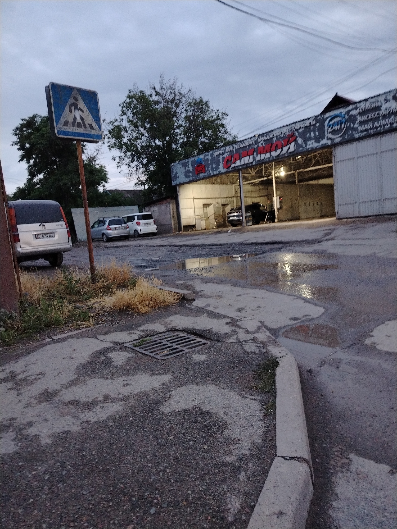 The image depicts an outdoor scene in Bishkek City, Kyrgyzstan, featuring a car wash facility with a sign that reads "CAR WASH" in large red letters. In the foreground, a pedestrian crossing sign is visible on a pole, indicating a crosswalk. Several vehicles are parked near the car wash, and the ground appears wet, suggesting recent rain or cleaning activities. The sky is overcast, contributing to a dim, early evening atmosphere. The overall scene captures a typical urban environment with commercial activity.