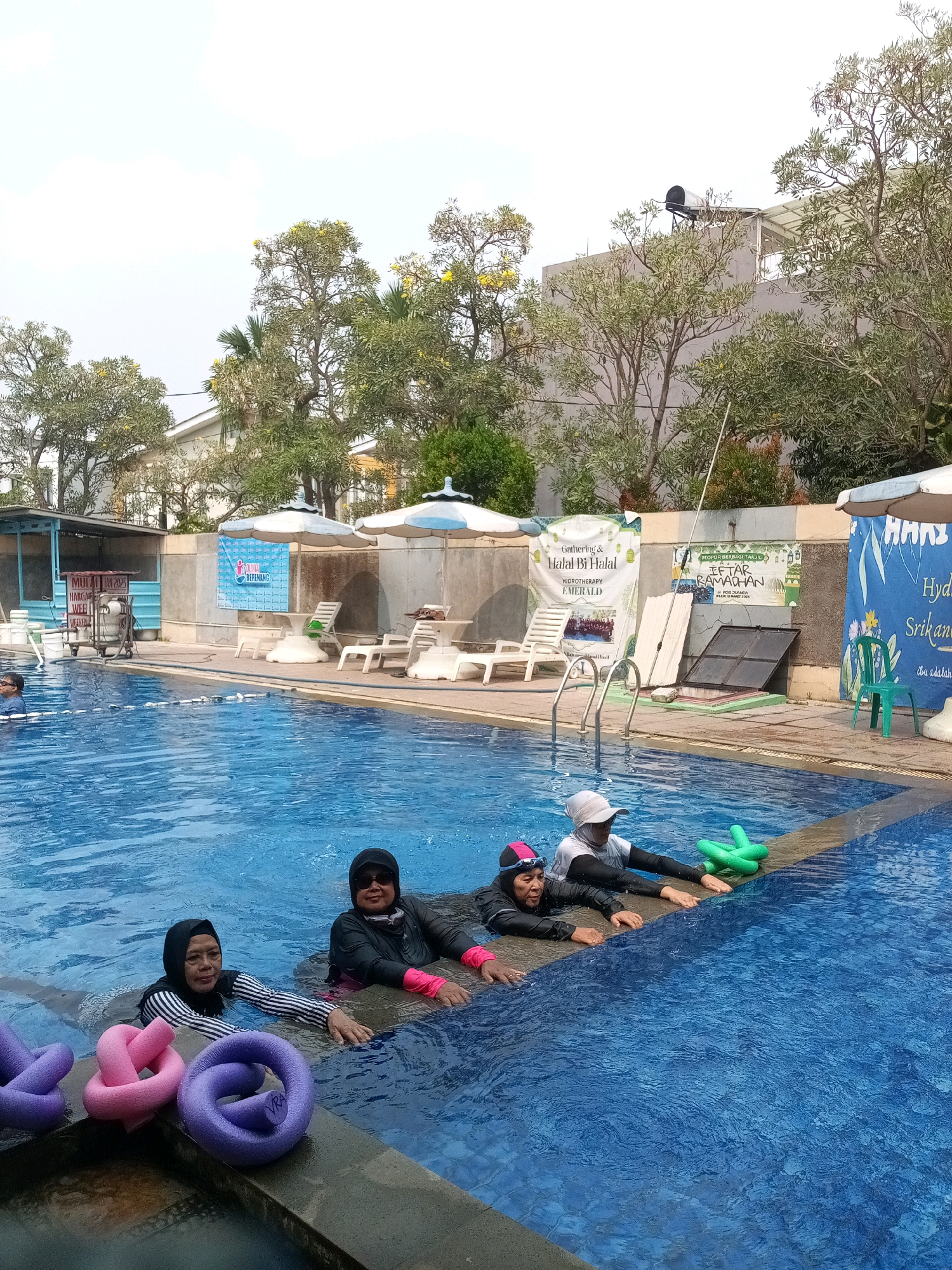 The image depicts an outdoor swimming pool area in Bekasi, Indonesia. In the foreground, four individuals are partially submerged in the bright blue water, leaning against the pool's edge. Three of them are women wearing modest swimwear, including headscarves and full-body suits. One woman in the foreground is wearing a white cap and black swimwear, leaning forward with her hands on the pool edge and holding green water noodles. Next to her, another woman in a black and white striped top and black headscarf is also resting her arms on the edge, looking towards the camera. To her right, a woman in black swimwear and sunglasses is also present. On the far left, partially obscured, another person is in the water. Scattered in the water near the pool edge are several colorful foam swimming aids, including purple and pink noodle shapes.
The background shows a paved area surrounding the pool with lounge chairs, tables, and large umbrellas providing shade. Various banners are displayed on a wall, one of which is blue with white lettering and appears to be an advertisement or announcement. Further back, there is a building with a light-colored facade, trees with green foliage and some yellow flowers, and a solar water heater on the roof of one structure. The overall scene suggests a recreational setting, possibly a public or private swimming facility.
