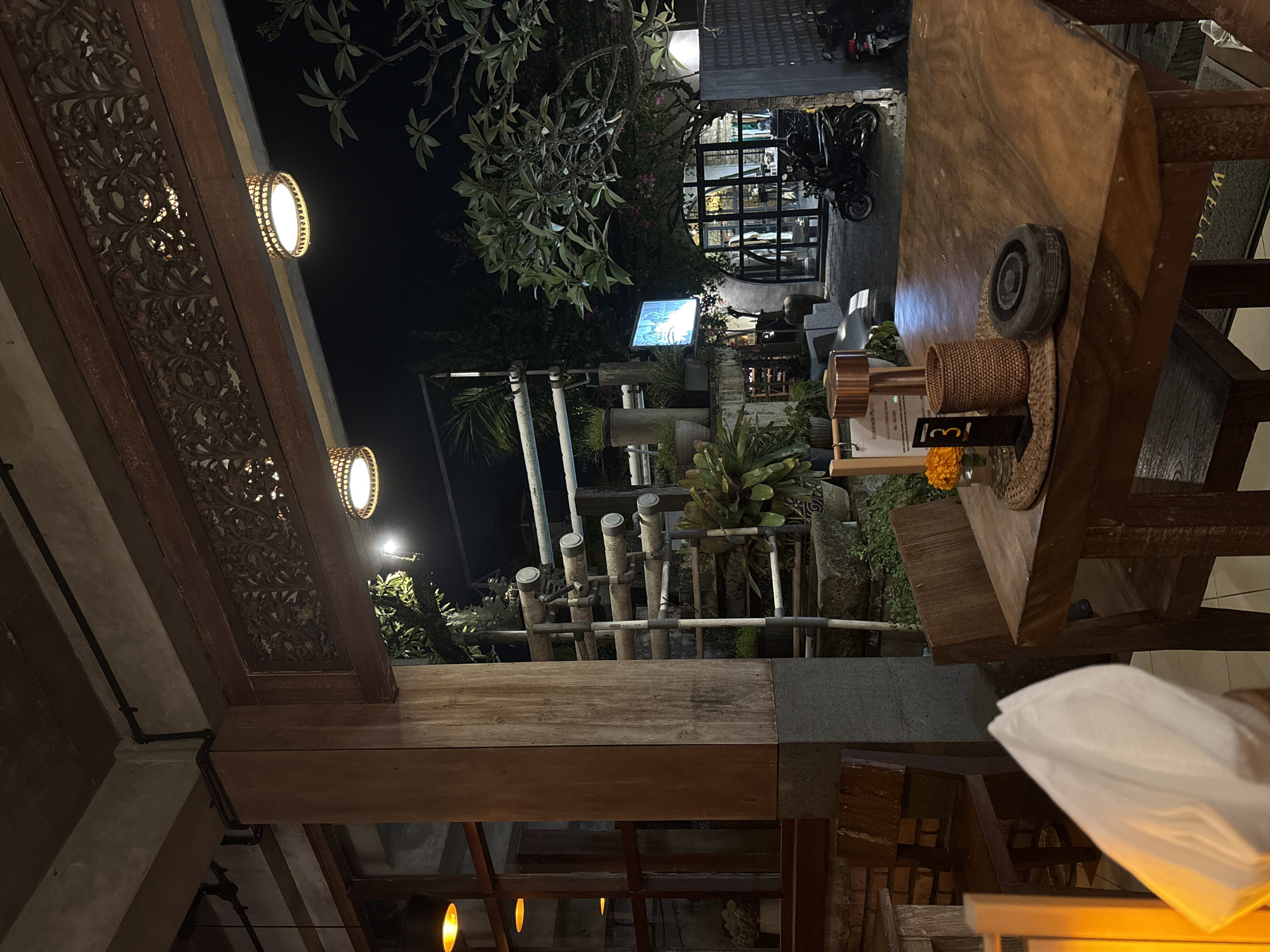 This image depicts an outdoor dining area in Ubud District, Indonesia, at night. The scene is partially illuminated by warm-toned lights attached to the building's wooden structure, showcasing intricate carvings. A wooden table dominates the foreground, bearing a decorative plate and various items. In the background, a building with glass walls is visible, alongside parked motorcycles and lush greenery. The outdoor area features decorative pipes and vegetation, adding to the tropical ambiance.