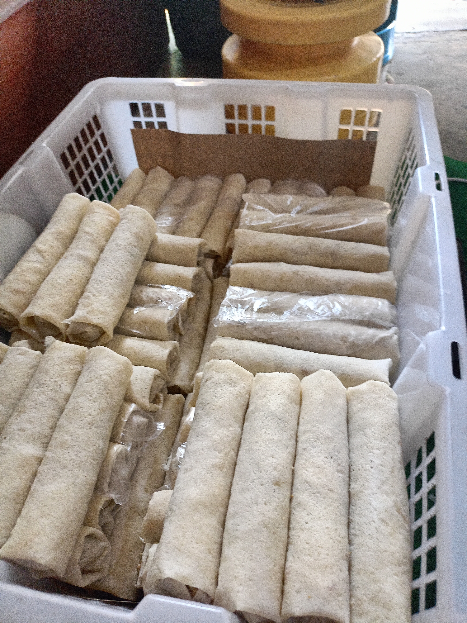 A white plastic basket is filled with uncooked spring rolls. The spring rolls are arranged in layers, with some wrapped in clear plastic. The basket has a grid pattern on the sides. In the background, there are yellow cylindrical objects and a dark blurry area. The scene appears to be an outdoor market stall in Malang, Indonesia.