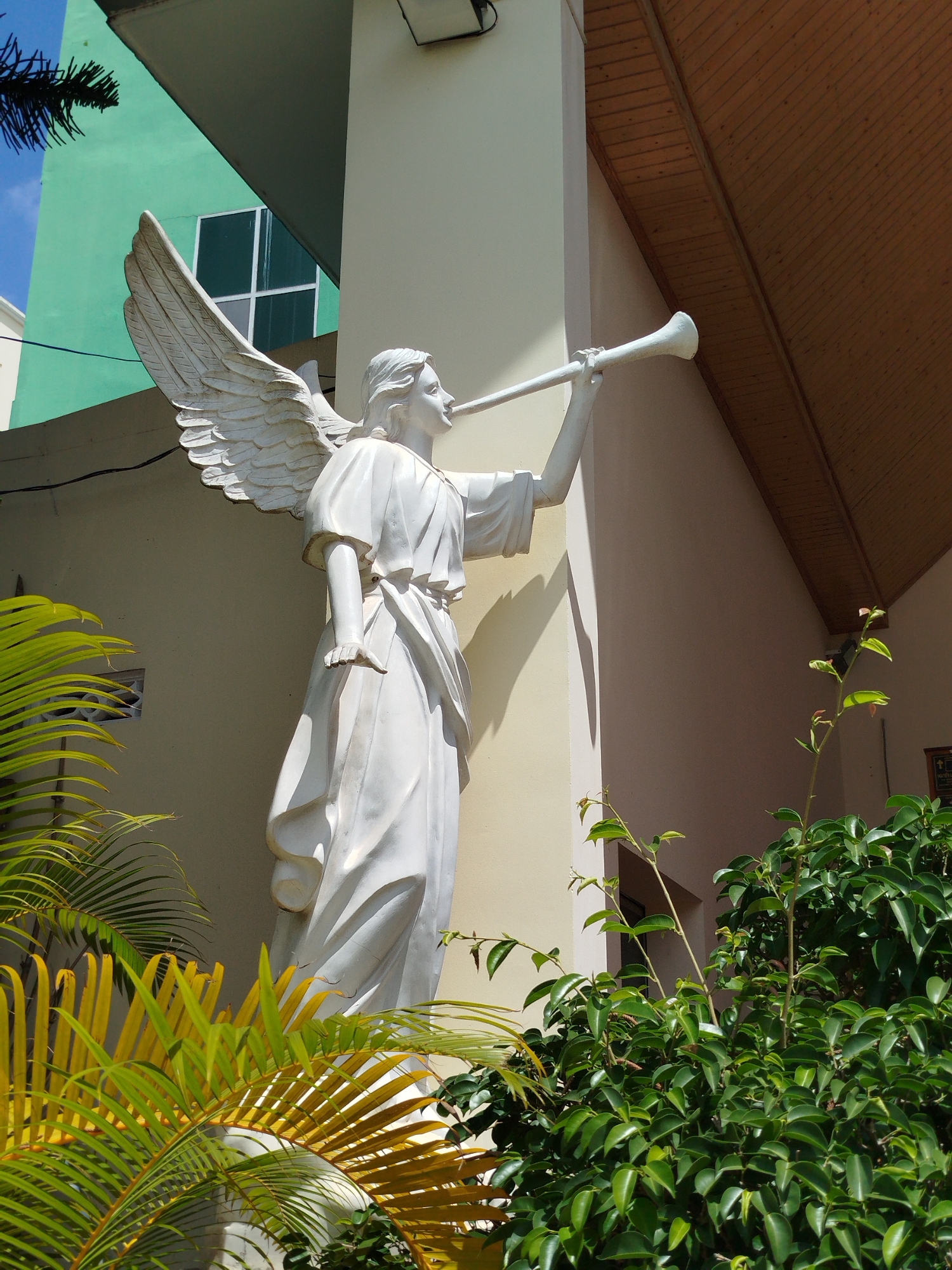 A statue of an angel blowing a trumpet stands outside a building in Pleiku Ward, Vietnam. The angel is white, and the building has a green and beige facade. Tropical plants surround the statue.