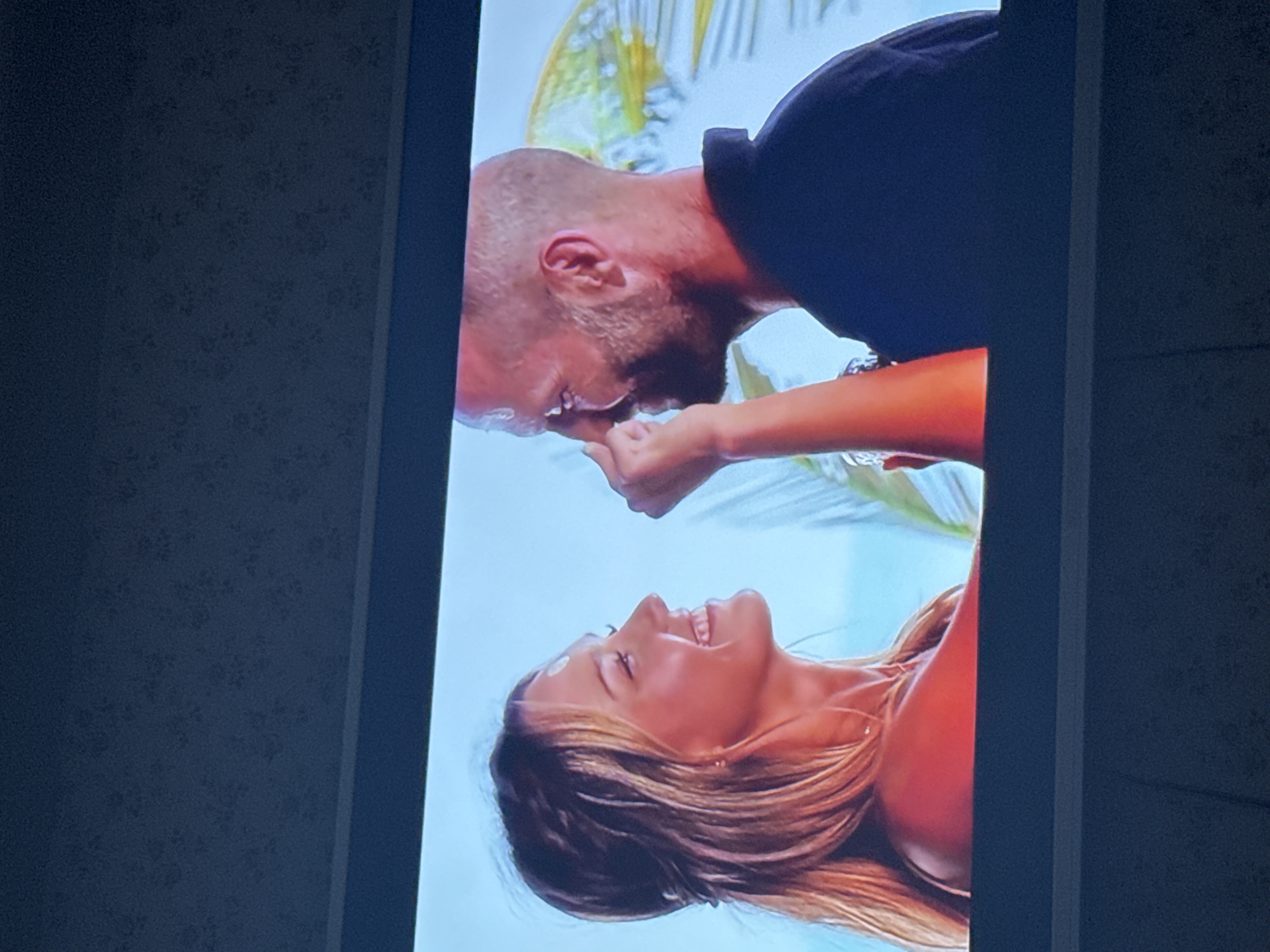 A television screen displays a couple facing each other. The man is bald, has a beard, and wears a black polo shirt. The woman has long, light brown hair and tanned skin. They are close to each other, possibly kissing or about to kiss. The background of the image on the screen appears to be a tropical or beach setting with palm trees. The television is set against a wall with a mottled, dark blue pattern. The location is not discernible from the image. Mytishchi, Russia is not visible in this image.