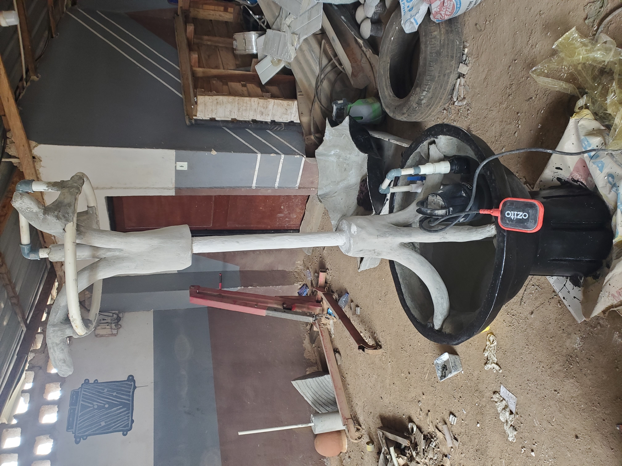 The image depicts an indoor space, likely a workshop or storage area, characterized by its rough construction and the presence of various objects. The floor is covered in dirt and sand, with scattered debris including wood scraps, plastic bags, and what appears to be a deflated tire. In the foreground, a black basin holds murky water, and a submersible pump with a red float switch is partially submerged. This basin is supported by a sculpted, light-colored structure resembling a pedestal with three outward-curving supports. Above this, a larger, circular, light-colored element is attached to a vertical pipe.
The background reveals a wall with a grey and brown color scheme, and a large, dark red door. To the left, a red engine hoist is positioned, and further left, a white pole stands upright in a concrete base. The ceiling is constructed of corrugated metal sheets supported by wooden beams, with gaps allowing sunlight to filter in. On the wall to the left, there's a stencil or mural of a black, rectangular object with white lines. The overall impression is one of an unfinished construction or a space used for storage and manual work, possibly in an area with limited resources or a developing infrastructure.
Based on the context of Jos, Nigeria, this scene could be found in a workshop area for artisans, a construction site, or a backyard storage space in a residential or commercial area of the city. The open-air feel of the roof suggests it might be a partially enclosed structure. The presence of the engine hoist and the sculpted elements could indicate that this is a space where construction, fabrication, or artistic creation is taking place.