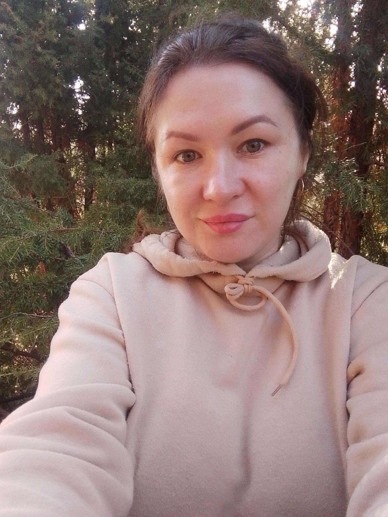 A selfie of a woman with dark hair pulled back in a loose bun, wearing a beige hoodie. She has fair skin, green eyes, and light pink lips. The background consists of green coniferous trees, possibly a park or wooded area. The lighting suggests it is daytime. There is no specific information to confirm that this is Прикамский, Russia.