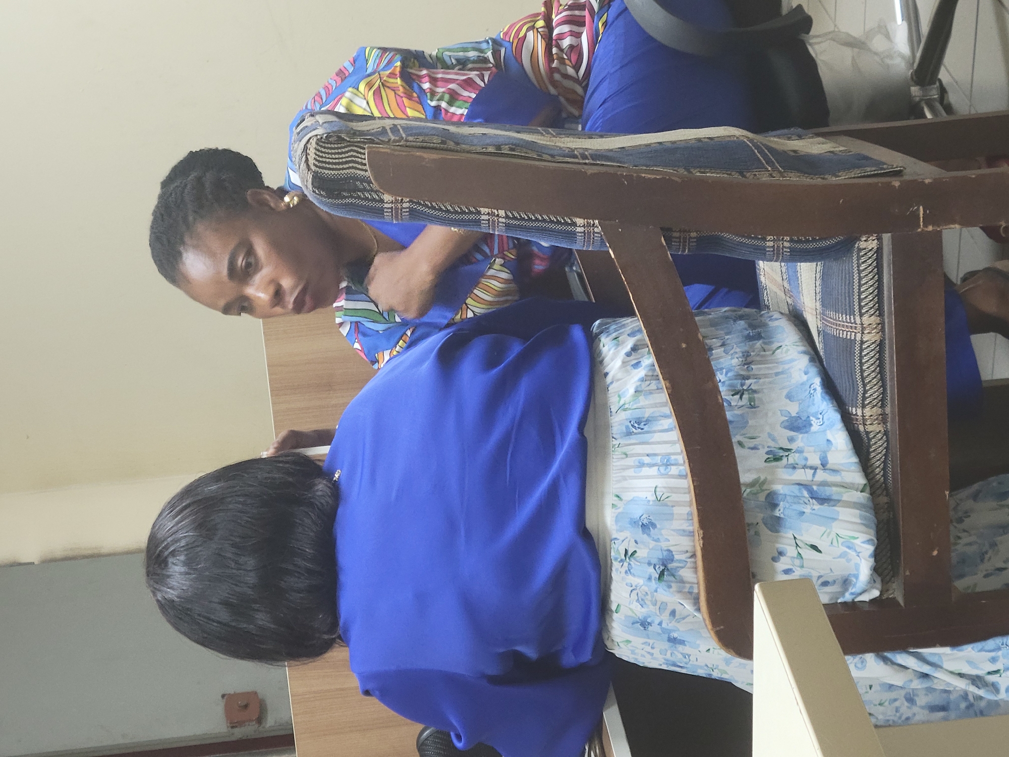 A woman with dark, braided hair is seated, leaning over a person seated in a wooden chair with blue and floral patterned upholstery. The woman is wearing a colorful patterned top and blue pants. The person in the chair is wearing a blue top. The location appears to be indoors. The city of Abuja, Nigeria is not discernible from the image.