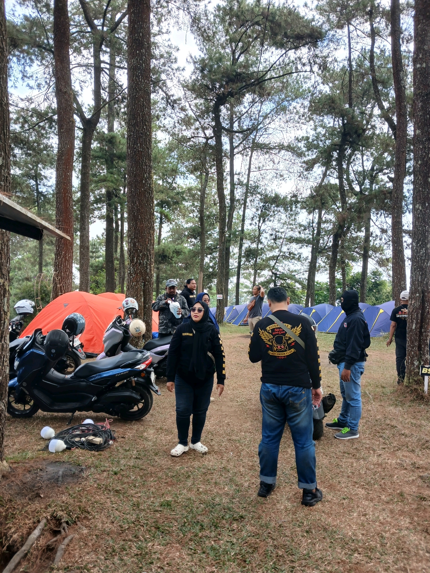 This is a medium shot taken outdoors in a wooded area, likely a campground, with tall pine trees dominating the background and foreground. In the middle ground, several tents are set up, one a bright orange and the others a uniform blue. A group of people, mostly men, are gathered around motorcycles and camping equipment. One man in the foreground wears a black long-sleeved shirt with a yellow emblem on the back, and blue jeans. He is facing away from the camera, with his back towards the viewer. Next to him, a woman in a black headscarf and jacket walks past. A blue motorcycle is parked in the foreground, and other motorcycles are visible in the background. A bundle of electrical cords and two white spheres, possibly lights, are on the ground near the blue motorcycle. The ground is covered in dry grass and fallen pine needles. The sky is overcast. The overall scene suggests a group of motorcyclists on a camping trip in a natural setting. The location is identified as Berjo, Indonesia.