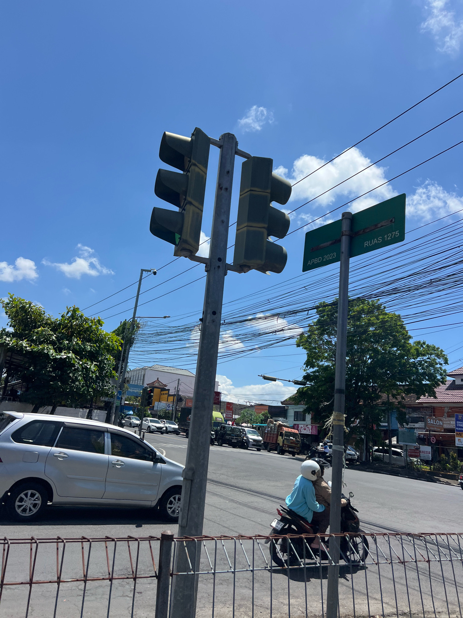 This image depicts a street scene in Denpasar, Indonesia, likely an intersection, on a sunny day with a bright blue sky and scattered clouds. A tall traffic light pole stands prominently in the foreground, supporting two sets of traffic signals. One set is facing the camera, with the green light illuminated. To the right of the traffic light pole is a green street sign that reads "APBD 2023" and "RUAS 1275".
On the road, several vehicles are visible. A silver minivan is parked or moving slowly in the left lane. A motorcycle carrying two individuals, one wearing a light blue shirt and white helmet and the other a brown jacket, is traveling on the road. In the background, other vehicles, including a truck and several cars, are present. Commercial buildings line the street in the distance. The scene is framed by a metal fence in the lower portion of the image. Overhead power lines crisscross the sky, a common sight in Indonesian cities. The overall impression is of a busy urban environment under a clear sky.