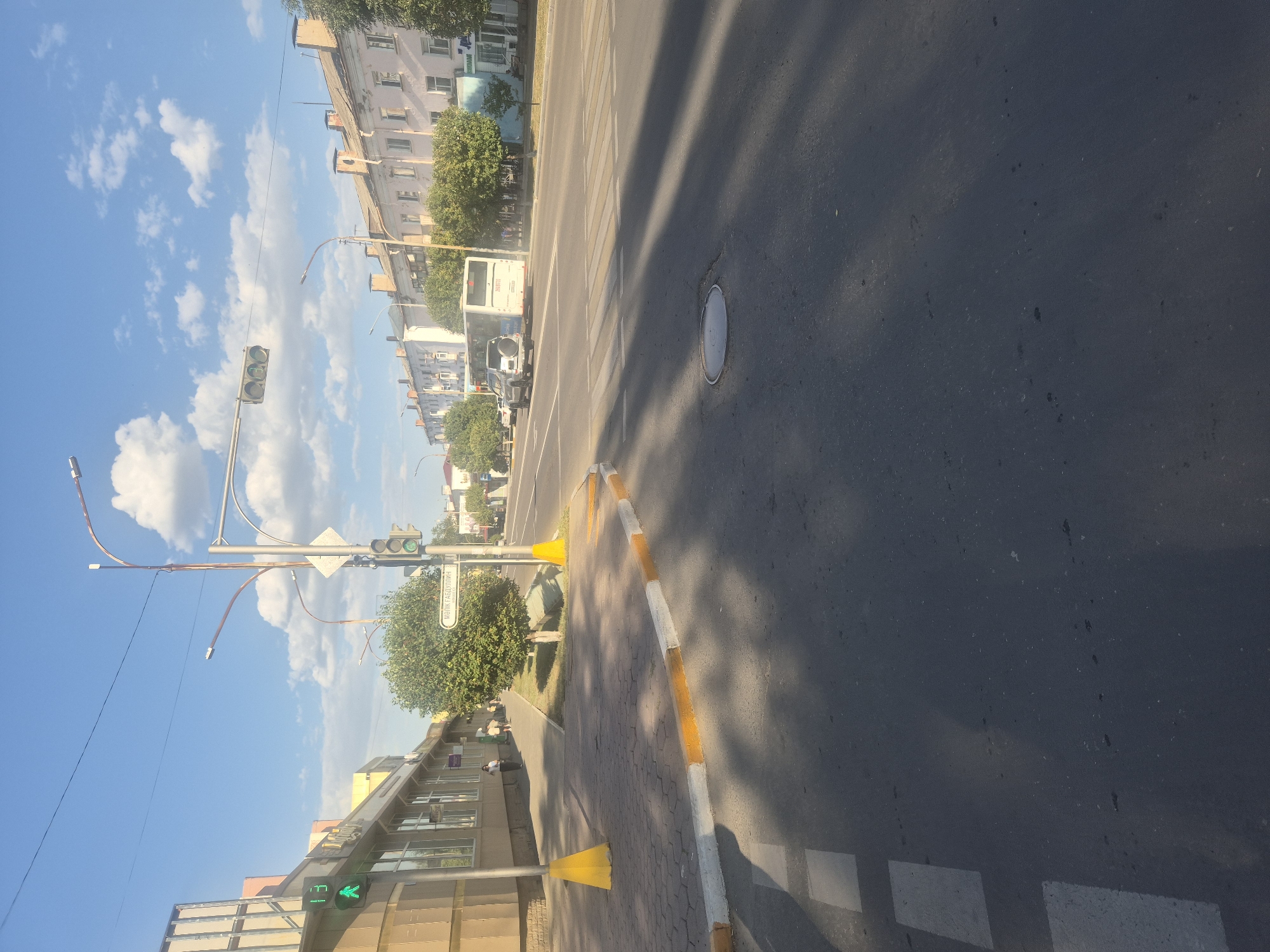 The image depicts a street scene in Kokshetau, Kazakhstan. In the foreground, a paved road with visible pedestrian crossing markings and a manhole cover. The right side of the road is bordered by a curb painted with alternating yellow and white stripes, suggesting it's a traffic island or a boundary. To the left of the road, a sidewalk is visible, with a pedestrian signal showing a green walking figure and the number "13" displayed. Adjacent to the sidewalk are buildings, one with a tan facade and signage. Several trees are planted along the sidewalk and beyond. Across the street, a row of light-colored buildings with pitched roofs lines the horizon, suggesting a residential or commercial area. Traffic signals are present at the intersection. A white bus and a silver SUV are driving on the road. The sky is blue with scattered white clouds. The overall impression is a sunny day in an urban environment.