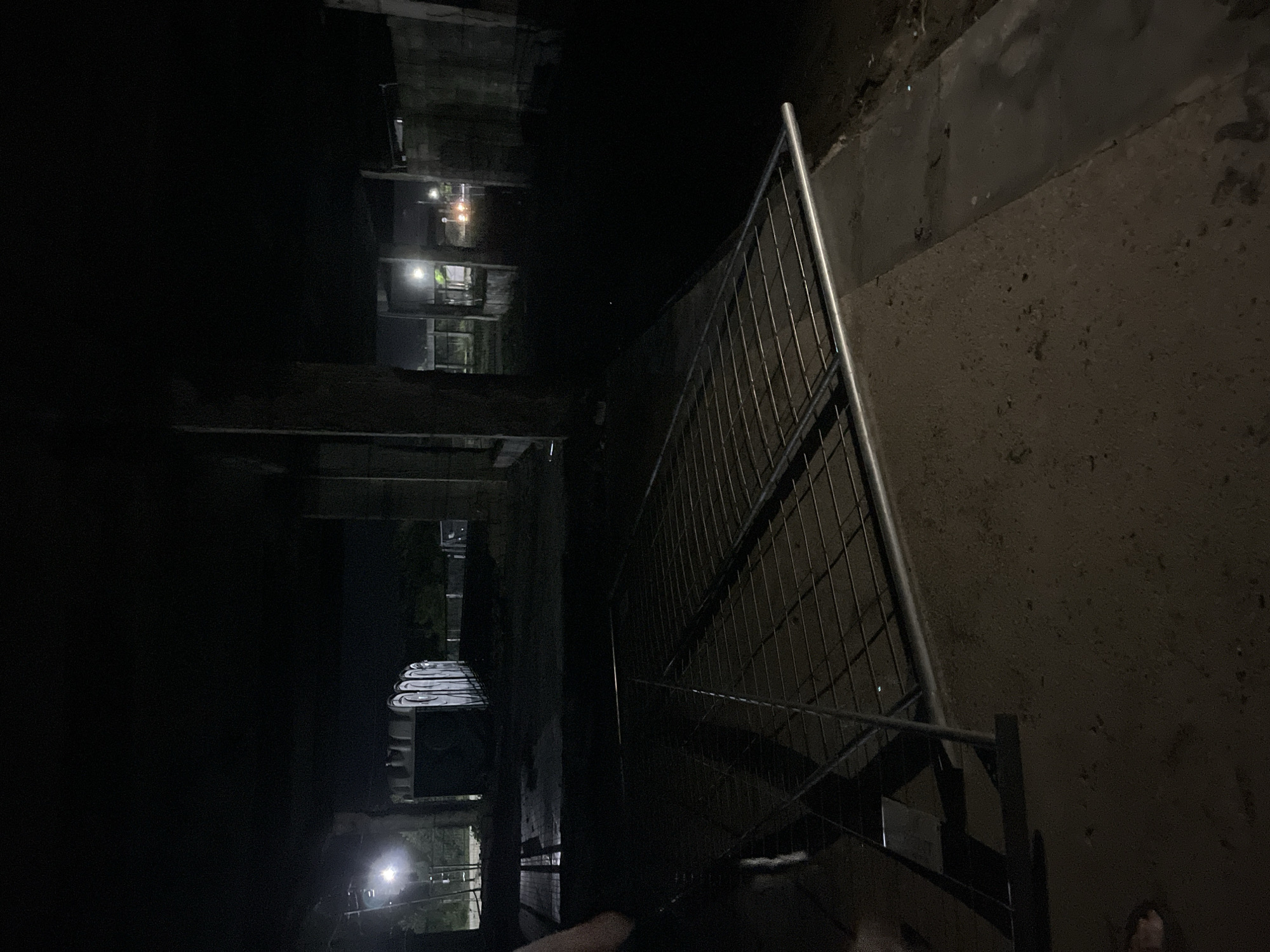 The image captures a dimly lit outdoor scene at night, likely in the city of Poti, Georgia. In the foreground, a metal grate or panel lies on the ground, angled across the frame. The surface beneath the grate appears to be concrete or a similar hard, uneven material. In the mid-ground, there are several portable toilets lined up. Beyond the toilets, the environment suggests an area with some construction or unfinished structures, evidenced by concrete pillars and rough walls. Lights are visible in the distance, including streetlights and what appear to be vehicle headlights. The overall atmosphere is dark and somewhat desolate, with shadows playing a significant role in obscuring details.