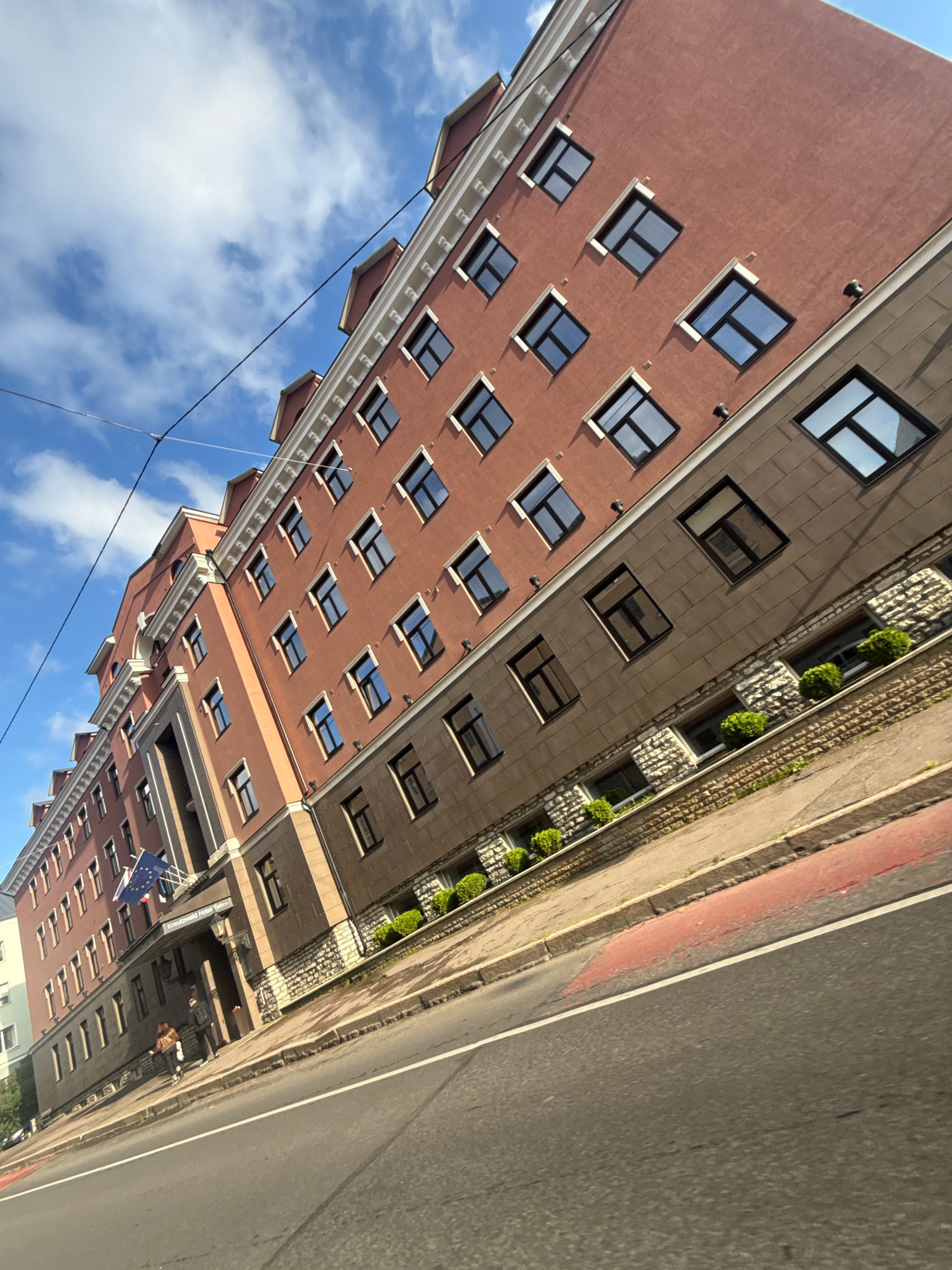 The image shows a long, multi-story building with a reddish-brown facade and a stone lower level, viewed from a low angle on a city street. The building has numerous windows, many of which appear to be dark or reflective. The architecture suggests it is a hotel or an institutional building. A sign above an entrance reads "Kreutzwald Hotel Tallinn". Two individuals are walking on the sidewalk in front of the hotel entrance. The street in the foreground is asphalt with red and white markings. The sky above is blue with white clouds, and power lines are visible. The scene is set in Tallinn, Estonia, specifically at the Kreutzwald Hotel.