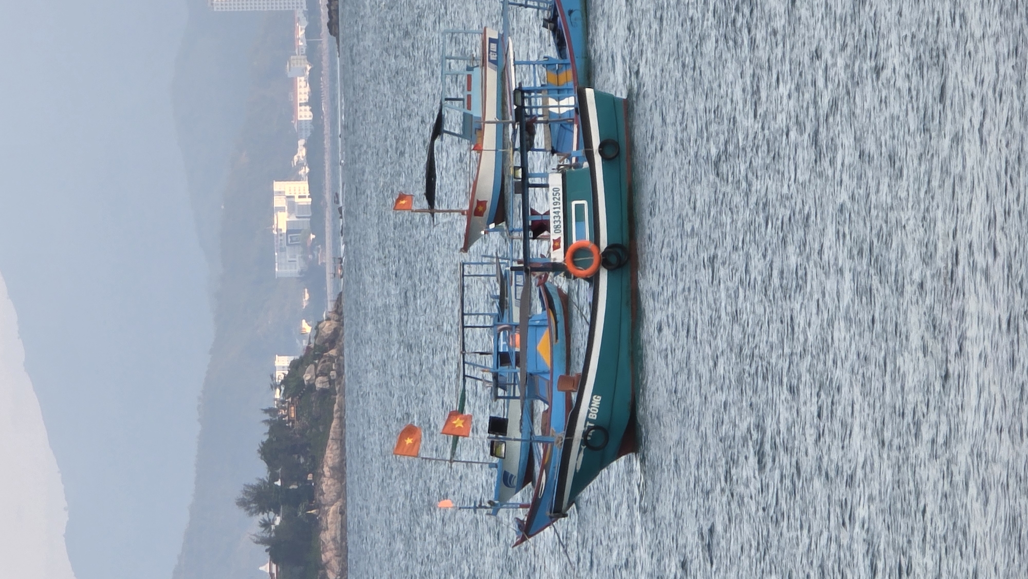 This image captures a serene coastal scene, likely in Bac Nha Trang Ward, Vietnam, showcasing several traditional Vietnamese boats on calm waters against a hazy mountainous backdrop.
In the foreground, a cluster of fishing or passenger boats is visible, gently floating on the lightly rippled sea. The most prominent boat, positioned towards the right, is a long, narrow vessel with a distinctive teal-green hull accented by a white stripe. It features blue deck structures and railings, an orange lifebuoy hanging from its side, and a red Vietnamese flag with a yellow star fluttering from a pole. Text on its hull includes the name "T BONG" (partially visible) and a phone number "0833419250". Behind this, other similar boats are moored, one primarily blue with yellow accents, and another smaller white-hulled boat with the partially visible name "VIET AN". Multiple Vietnamese flags are visible across the boats, emphasizing the location's identity.
The water itself is a light blue-grey, covered in numerous small, parallel ripples, suggesting a gentle current and reflecting the soft, diffused light. The overall illumination indicates either early morning or late afternoon, with a gentle, diffused glow rather than harsh midday sun.
Beyond the boats, the coastline unfolds. The immediate shore consists of rocky formations interspersed with green trees. Further inland, several light-colored, multi-story buildings are visible, suggesting coastal development or resorts. These structures are scattered along the base of what appears to be a large mountain range, which dominates the background. The mountains are heavily obscured by atmospheric haze, appearing in muted blue and grey tones, creating a sense of depth and distance. The scene conveys a peaceful maritime atmosphere characteristic of a Vietnamese fishing or tourist port.