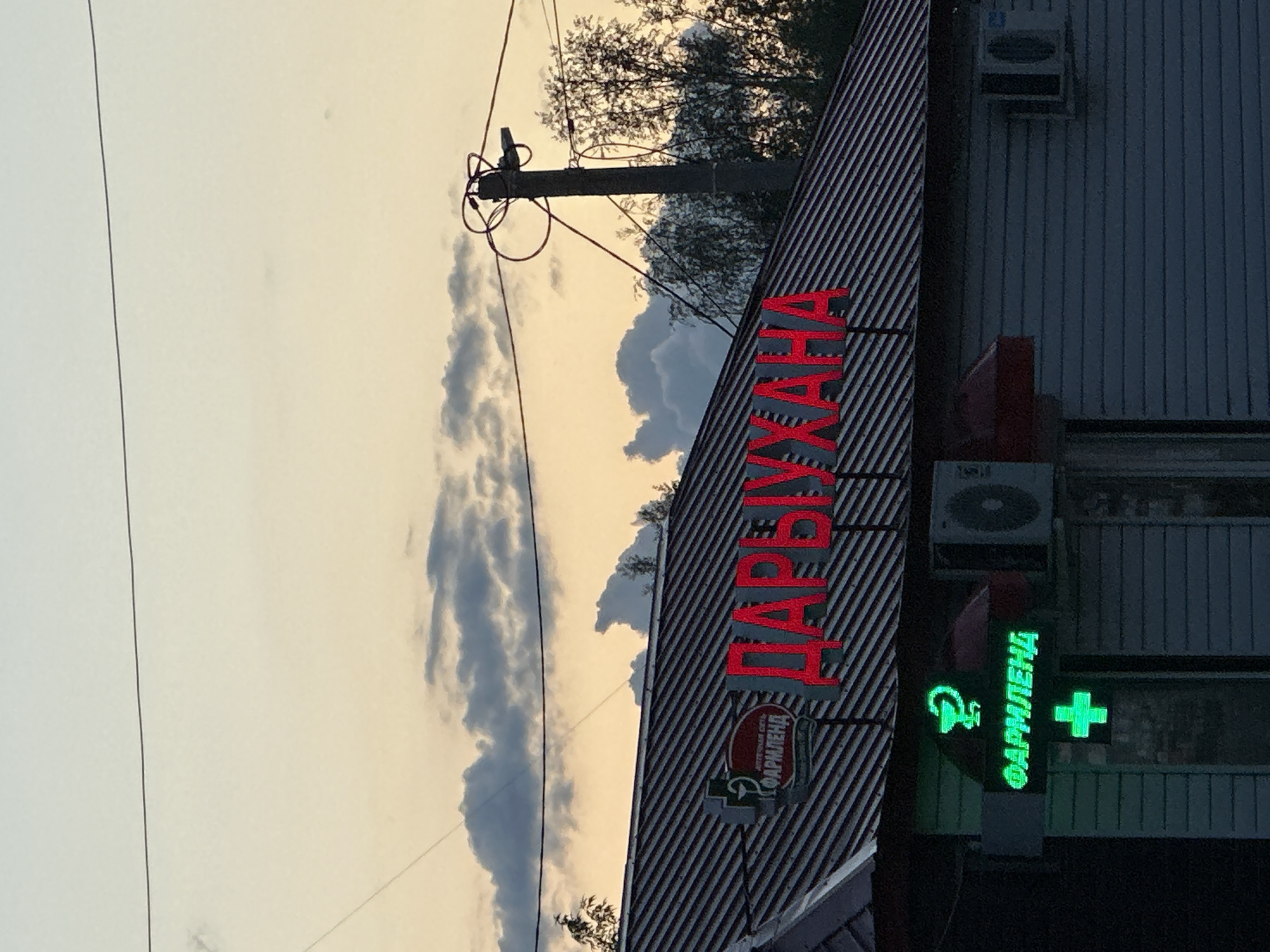 The image shows a commercial building, likely a pharmacy, judging by the neon sign in Cyrillic alphabet spelling "ФАРМЛЕНД" and a symbol with a bowl and a serpent associated with pharmacies. Above the pharmacy name is a larger sign in red neon, also in Cyrillic script, "ЦАРВІХАНА". The building has vertical siding and air conditioning units on the exterior. A utility pole with wires is visible against a partly cloudy sky. The image is rotated clockwise. The location is in Unknown, Unknown, based on the Cyrillic text on the signage. No people are visible.