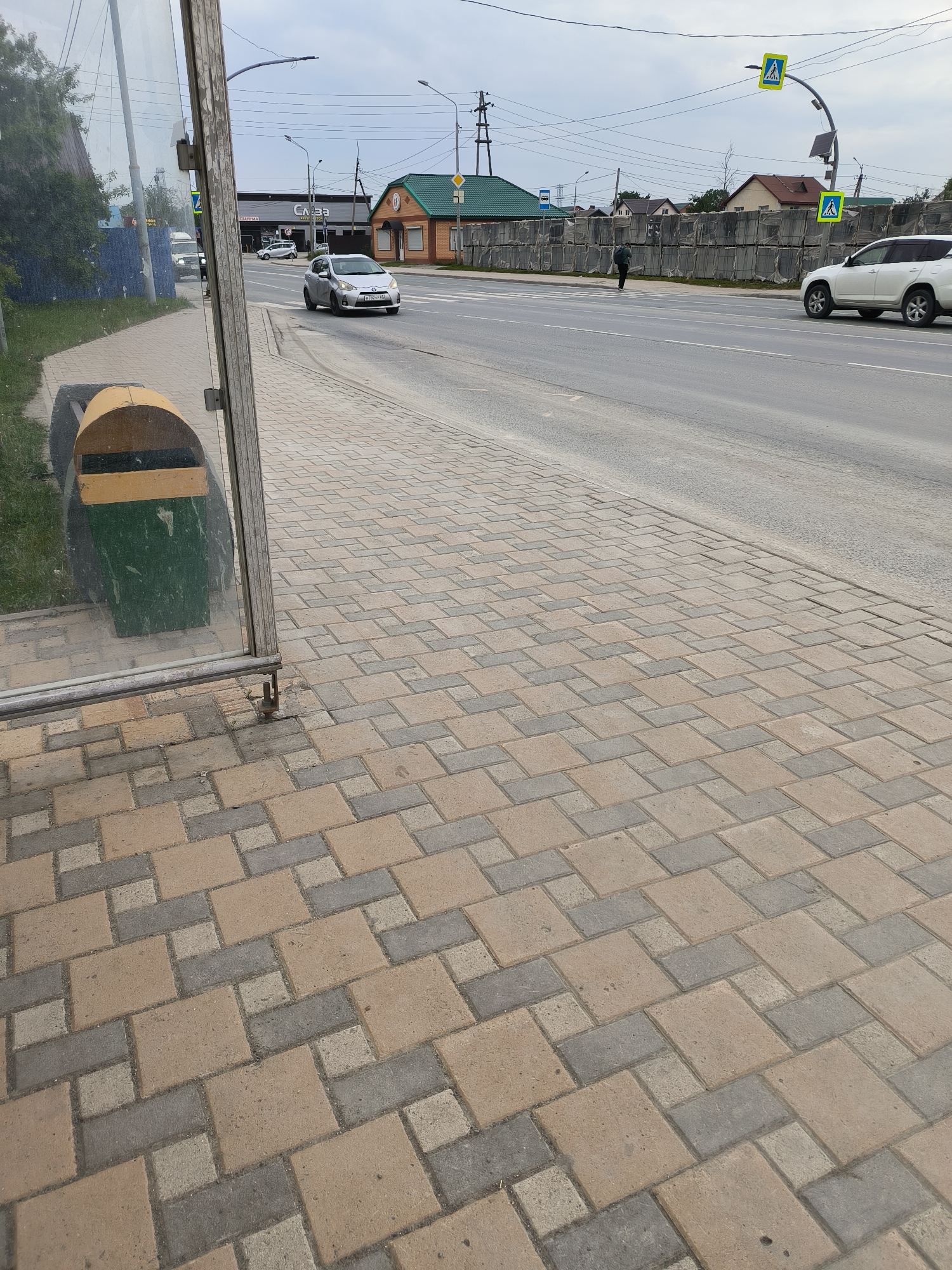 The image shows a street scene in Yuzhno-Sakhalinsk, Russia. A bus stop shelter with a glass panel and a trash can is in the foreground, partially obscuring the view of the street. The pavement is made of interlocking bricks in shades of tan and gray.
Across the street, there is a road with light traffic, including a silver sedan and a white SUV. Several buildings line the street, including a small, colorful building with a green roof and a long, gray building with "Слава" (Slava) written on it. Pedestrian crossing signs are visible, indicating a crosswalk. A lone individual is walking across the street in the distance.
The sky is overcast, suggesting a cloudy or possibly rainy day. The time of day appears to be daytime, likely afternoon. The overall impression is a typical urban or suburban street in Russia.